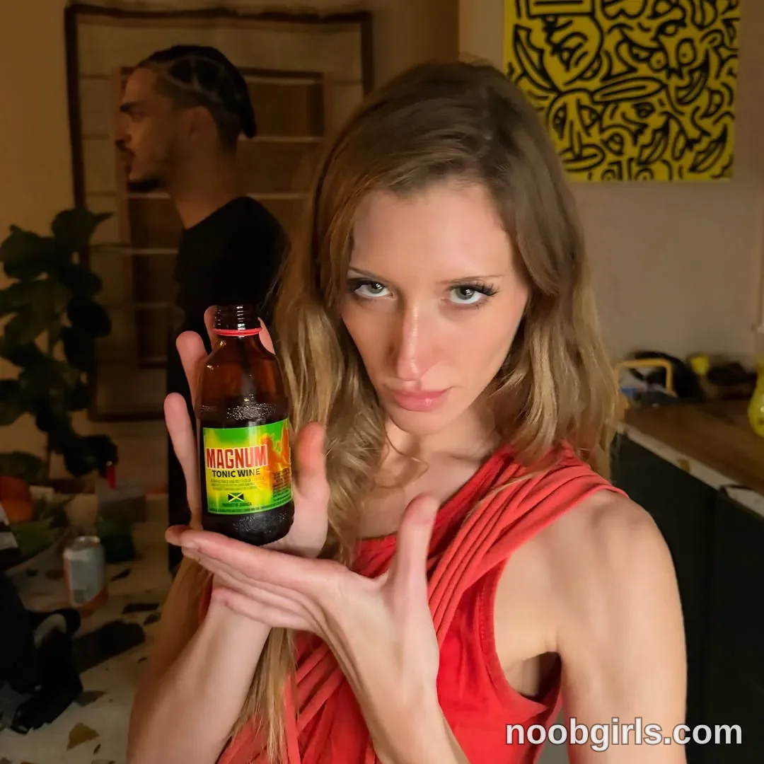 Addison Vodka Holding the bottle upright beside her face with one hand wearing Red sleeveless top with draped fabric in room
