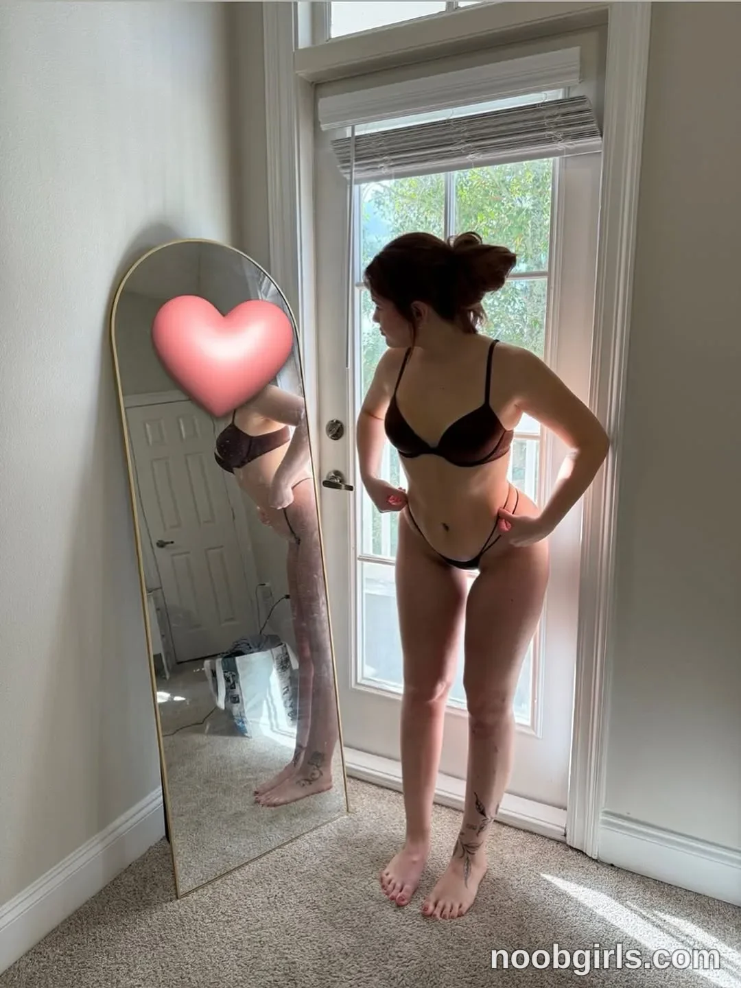 Ali Standing barefoot with her head turned toward the mirror wearing Black bra in a carpeted room beside a glass-paneled door