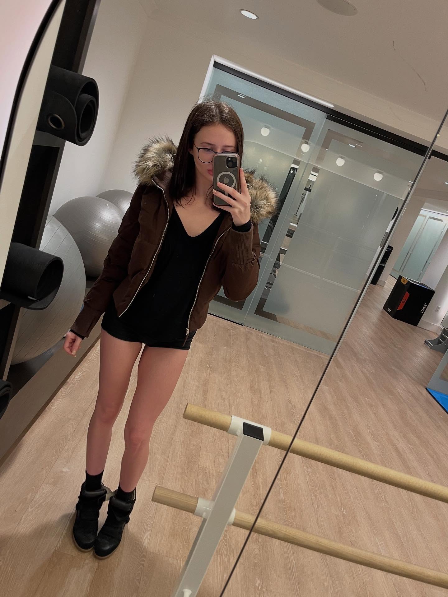 alyssapearls taking mirror selfie wearing brown jacket with fur-trimmed hood in fitness studio