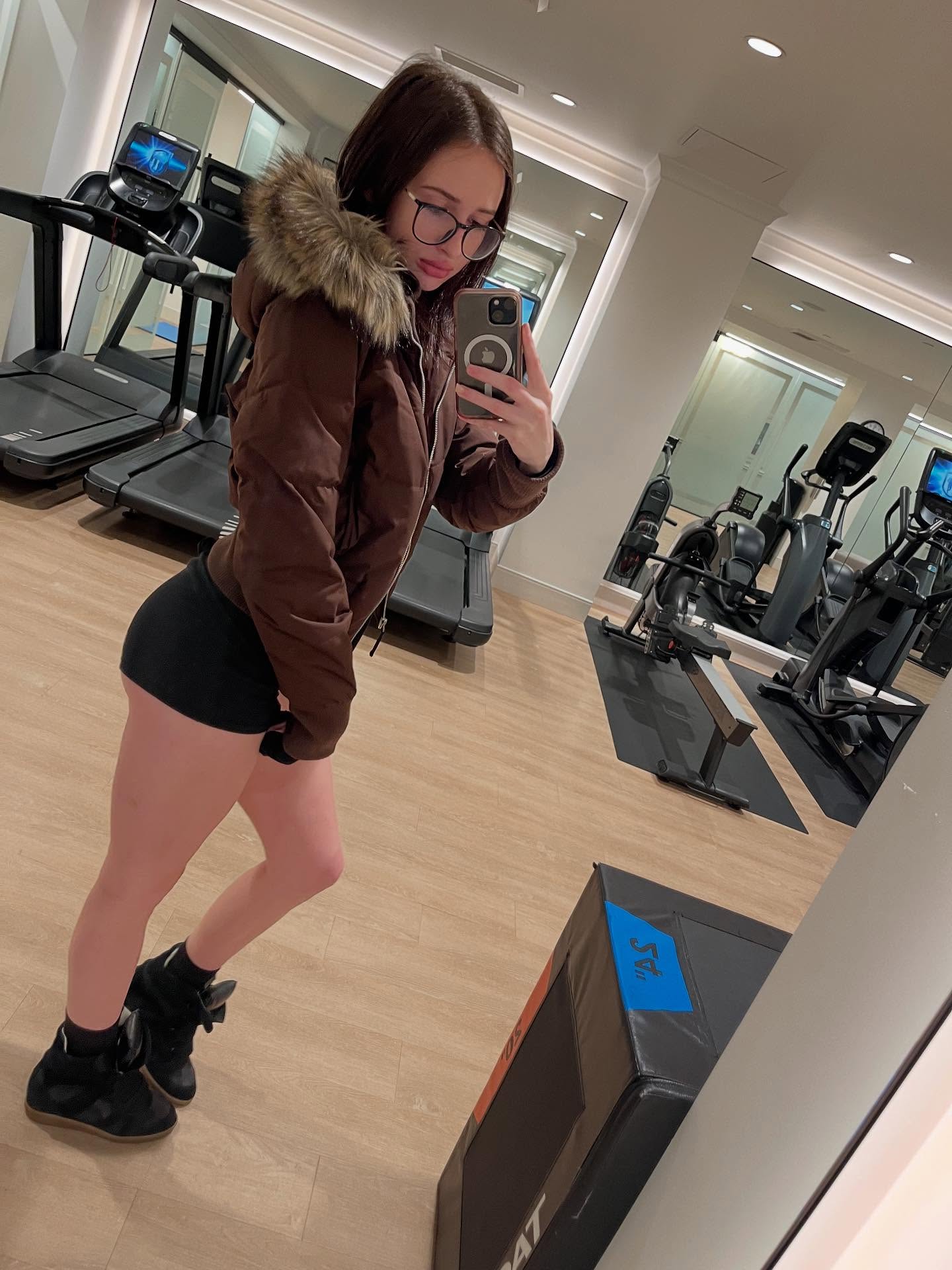 alyssapearls taking a mirror selfie wearing brown hooded puffer jacket with fur trim in gym