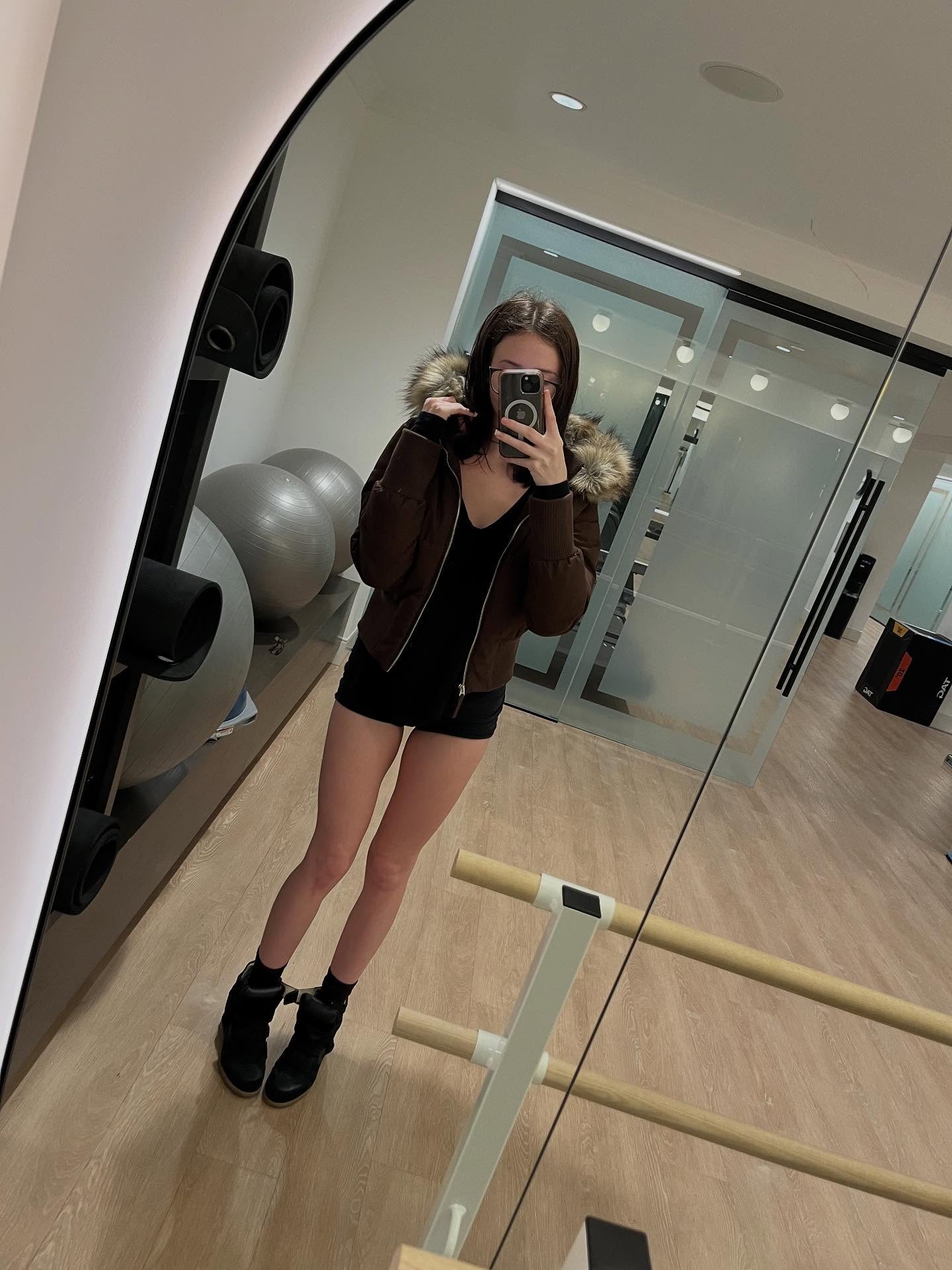 alyssapearls taking mirror selfie wearing brown jacket with fur hood in exercise studio