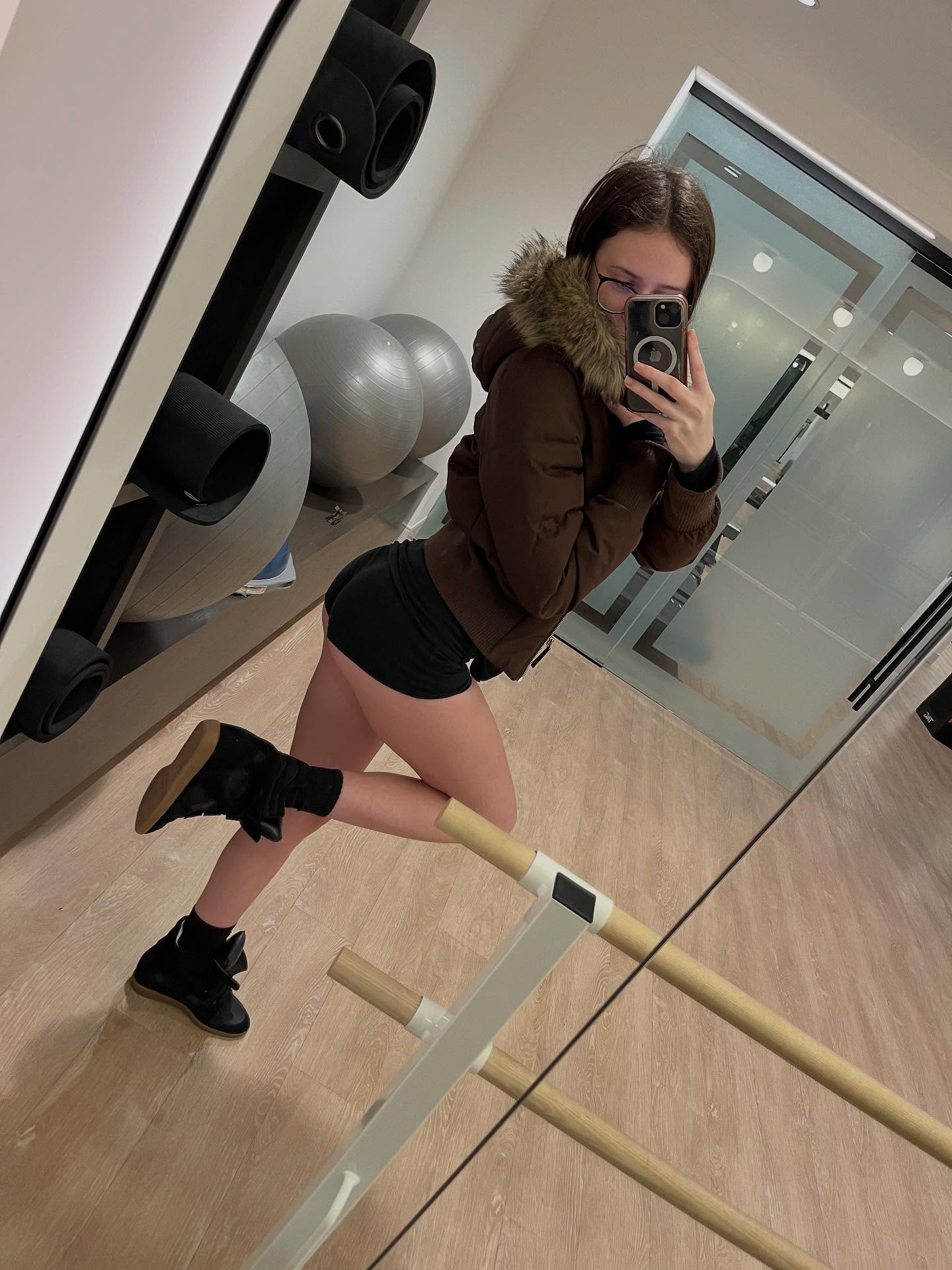 alyssapearls taking a mirror selfie wearing brown hooded jacket with fur trim in exercise studio