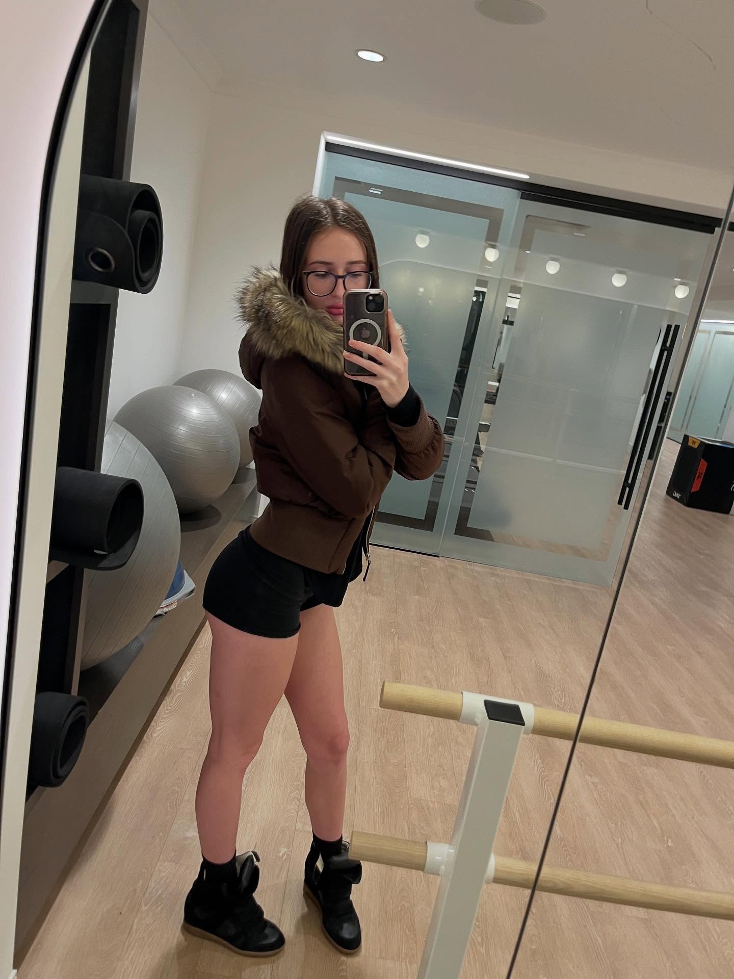 alyssapearls taking a mirror selfie wearing brown hooded jacket in gym room