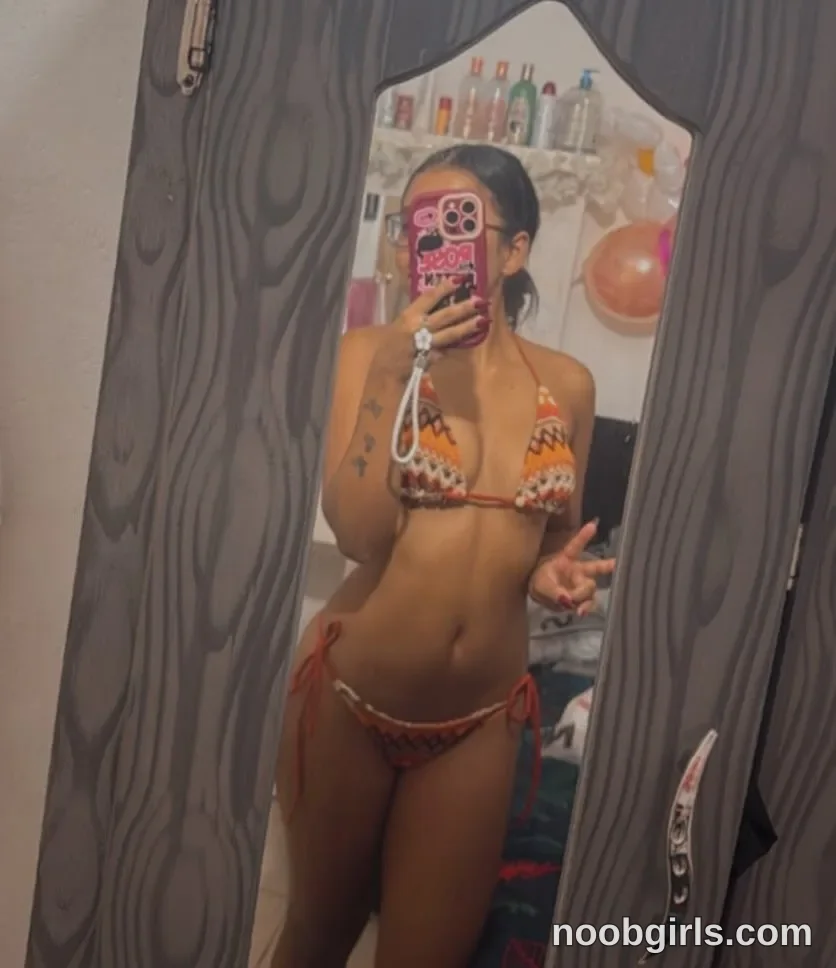Ana Now independent standing in front of the mirror wearing orange, white, and dark-patterned triangle bikini top in room