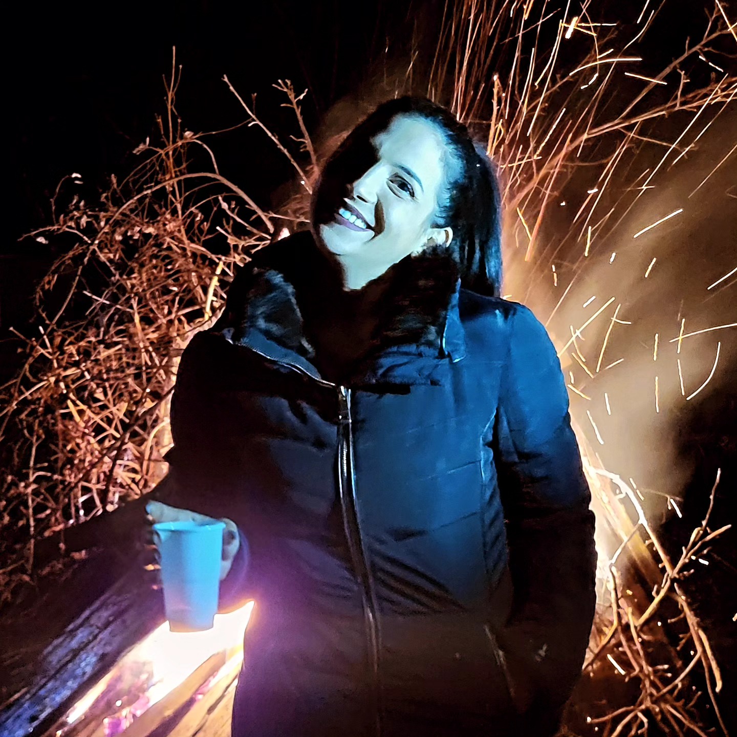 anaworld69 holding a cup wearing black winter jacket by bonfire