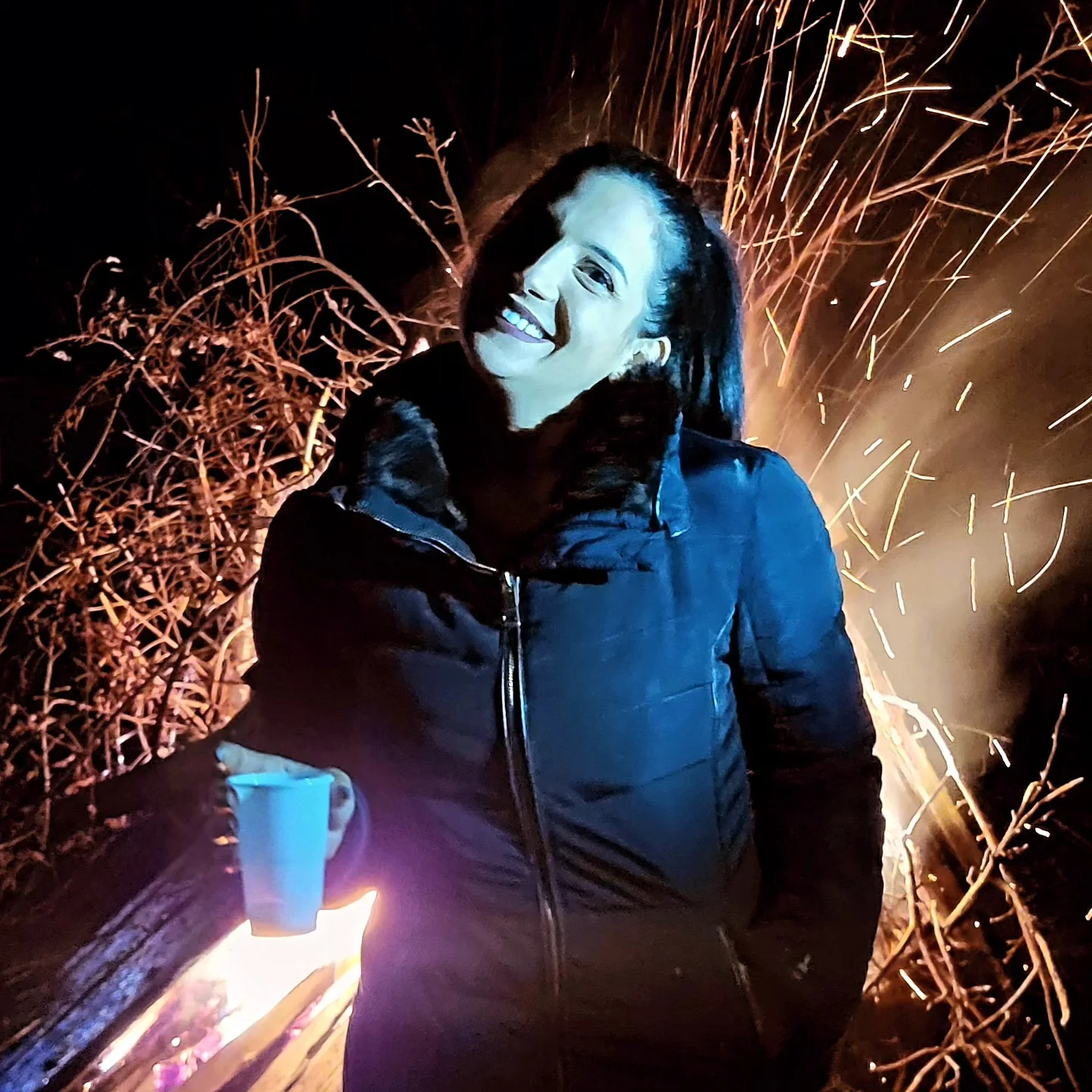 anaworld69 holding a cup wearing black puffer jacket with fur collar near bonfire