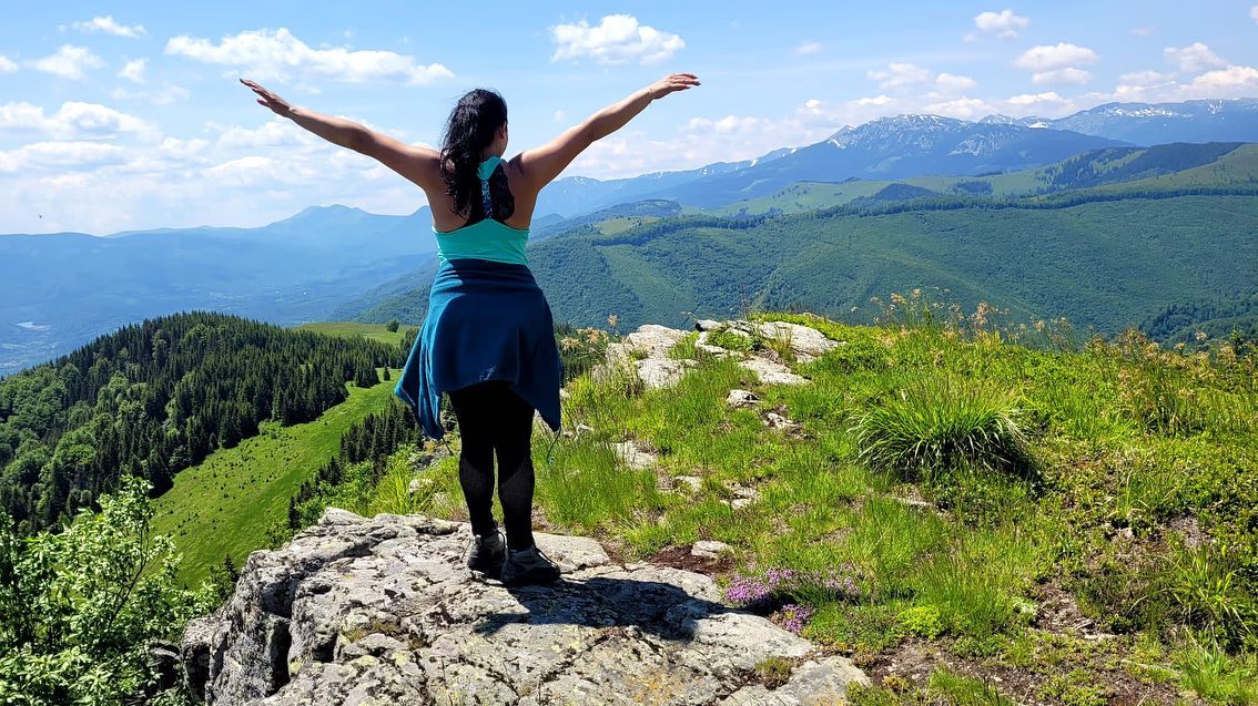 anaworld69 standing with arms outstretched wearing teal tank top in mountain summit