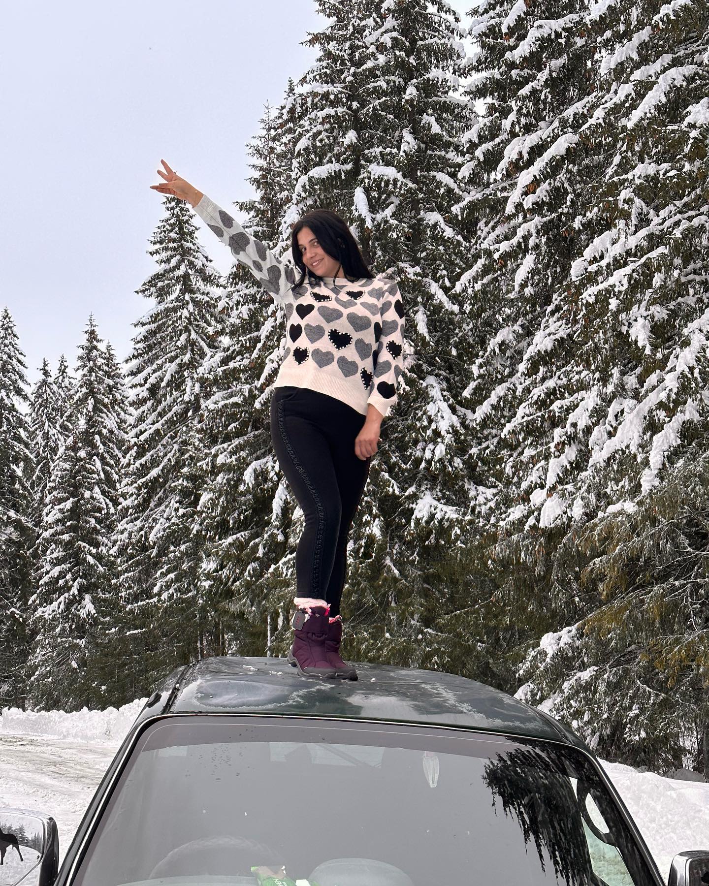 anaworld69 standing on vehicle roof wearing white patterned sweater in snowy pine forest