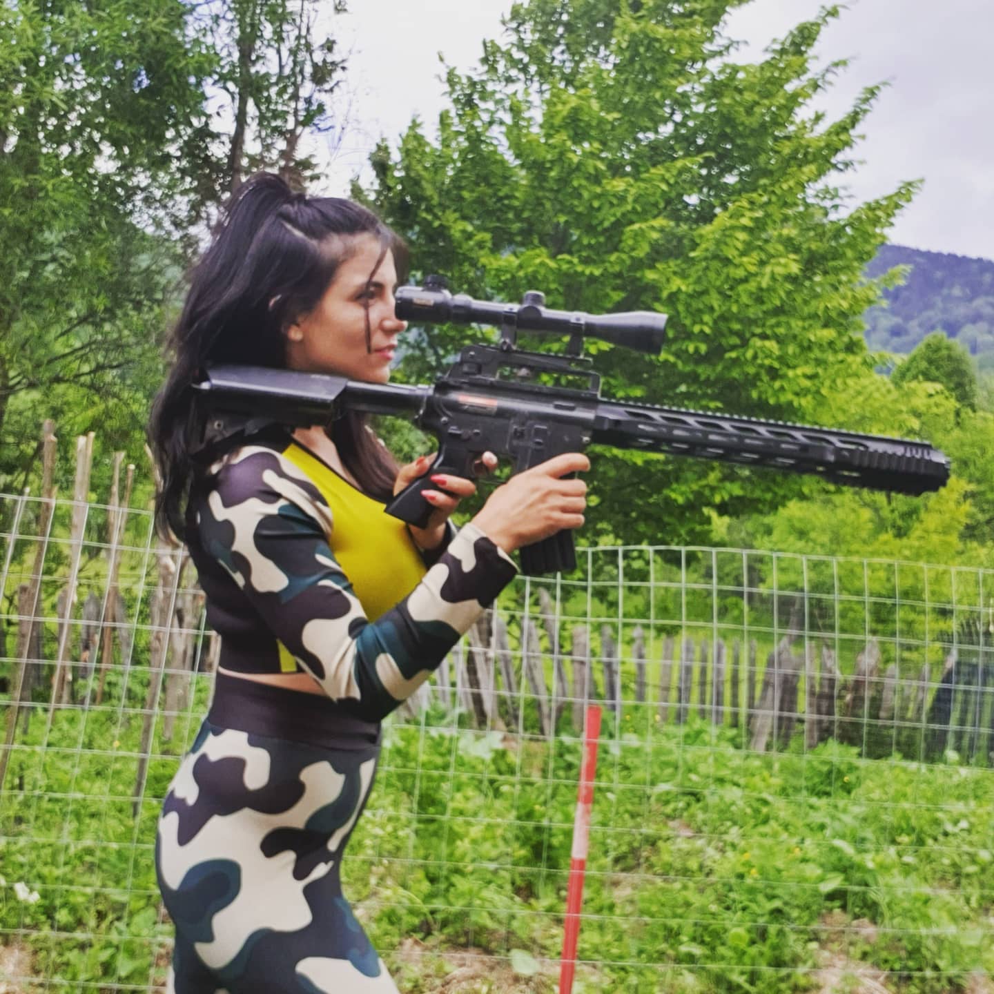 anaworld69 aiming a scoped rifle wearing camouflage and yellow top in yard