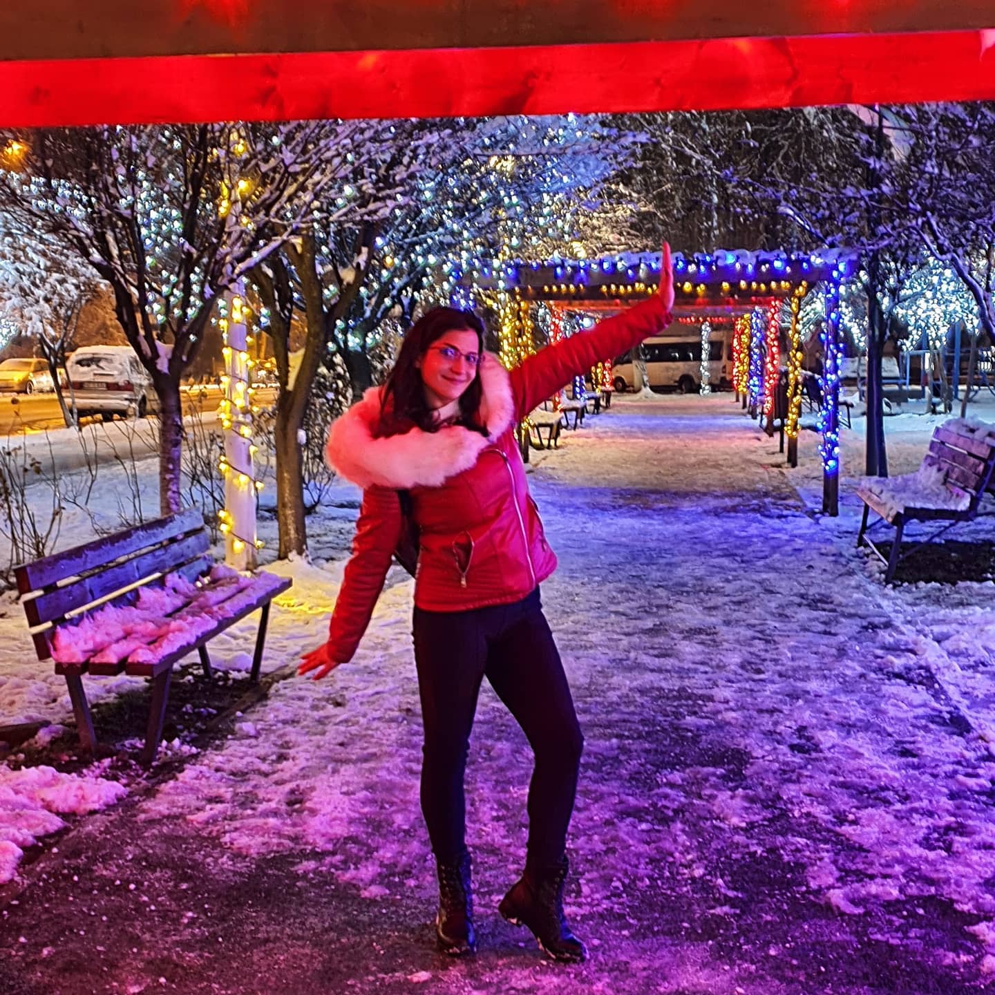 anaworld69 posing with arm raised wearing red jacket with fur trim in snowy illuminated walkway