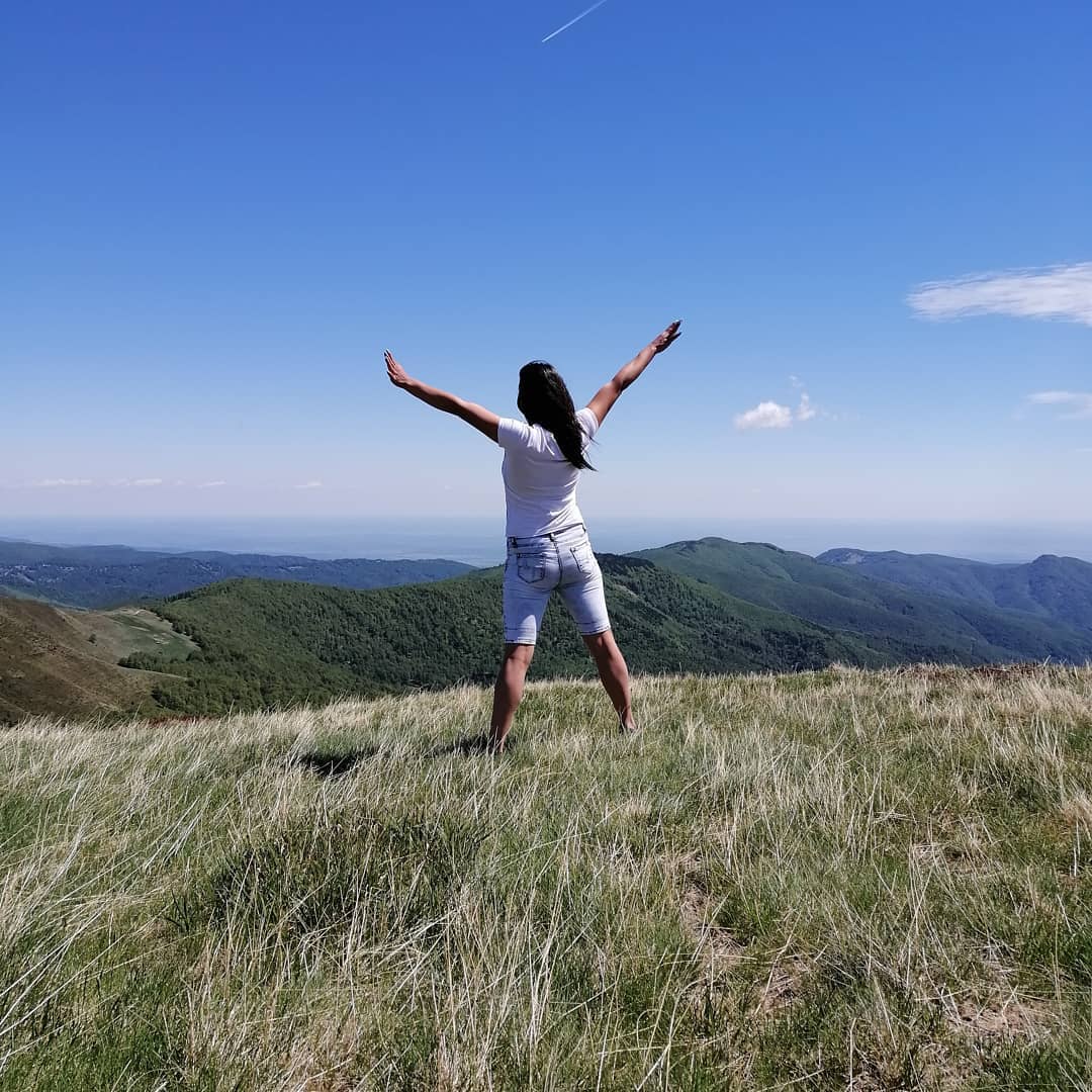 anaworld69 standing with arms raised wearing white t-shirt in grassy mountain summit