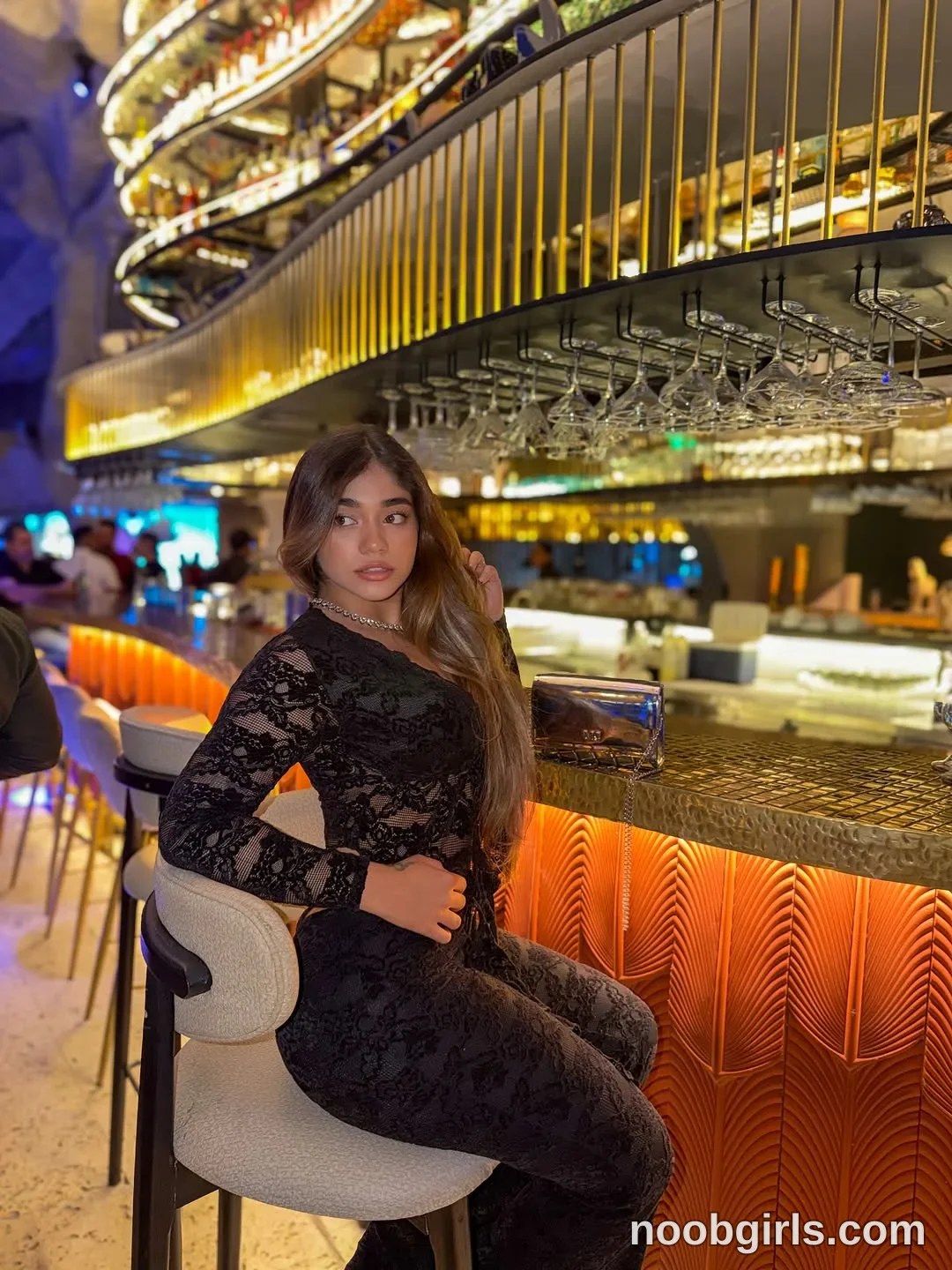 Ale sitting on a bar stool with one hand touching her hair wearing black sheer lace long-sleeved top in bar or lounge