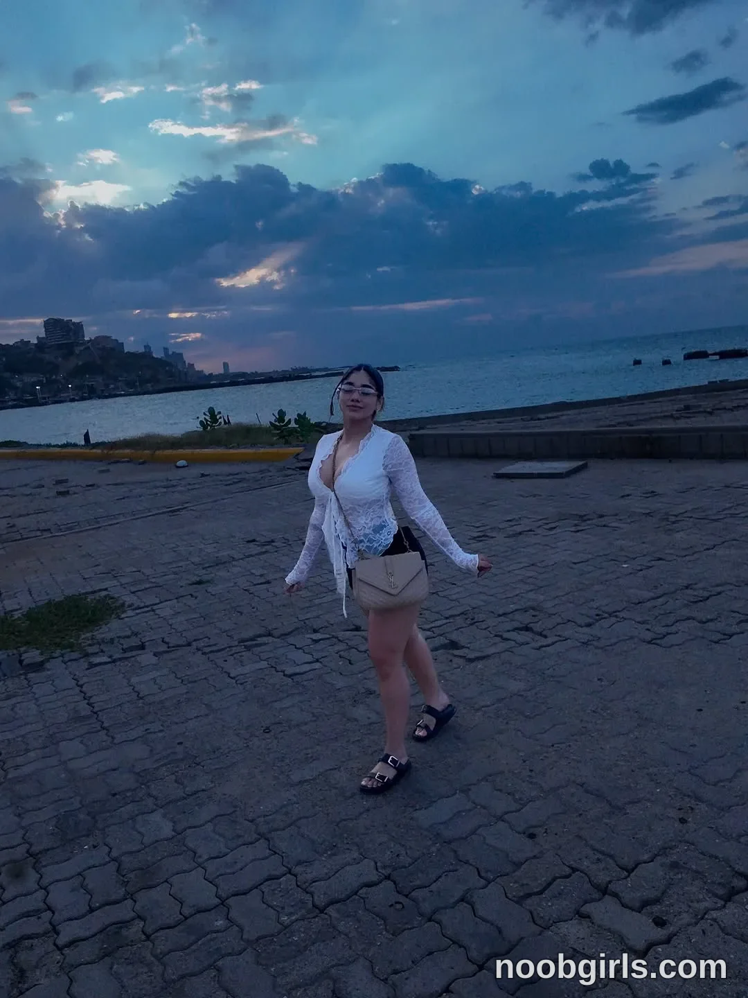 Ale walking or posing mid-stride on a paved stone surface wearing white long-sleeved sheer lace top