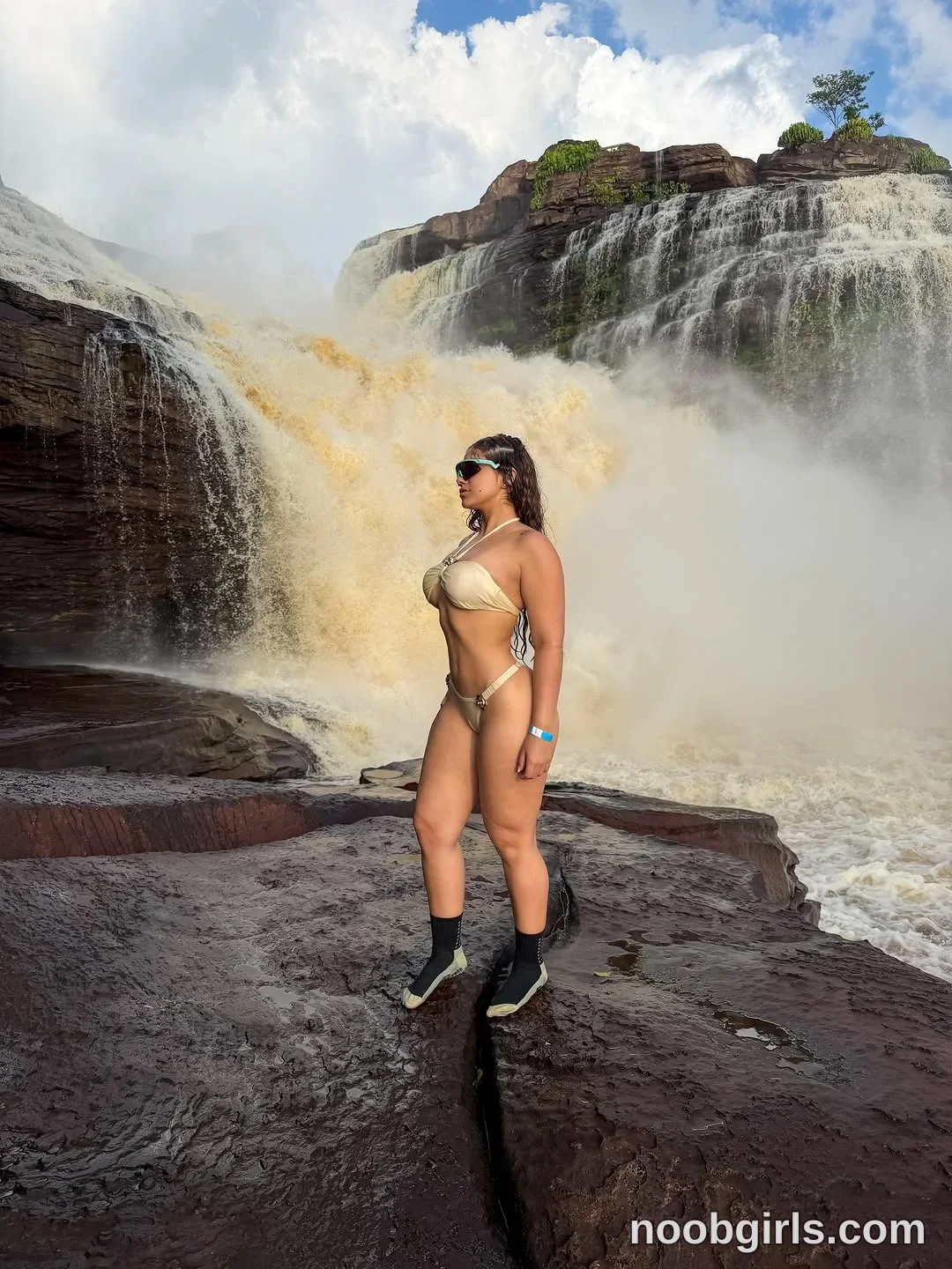 Ale standing on wet rocks facing sideways in front of a waterfall wearing metallic beige halter bikini top in waterfall