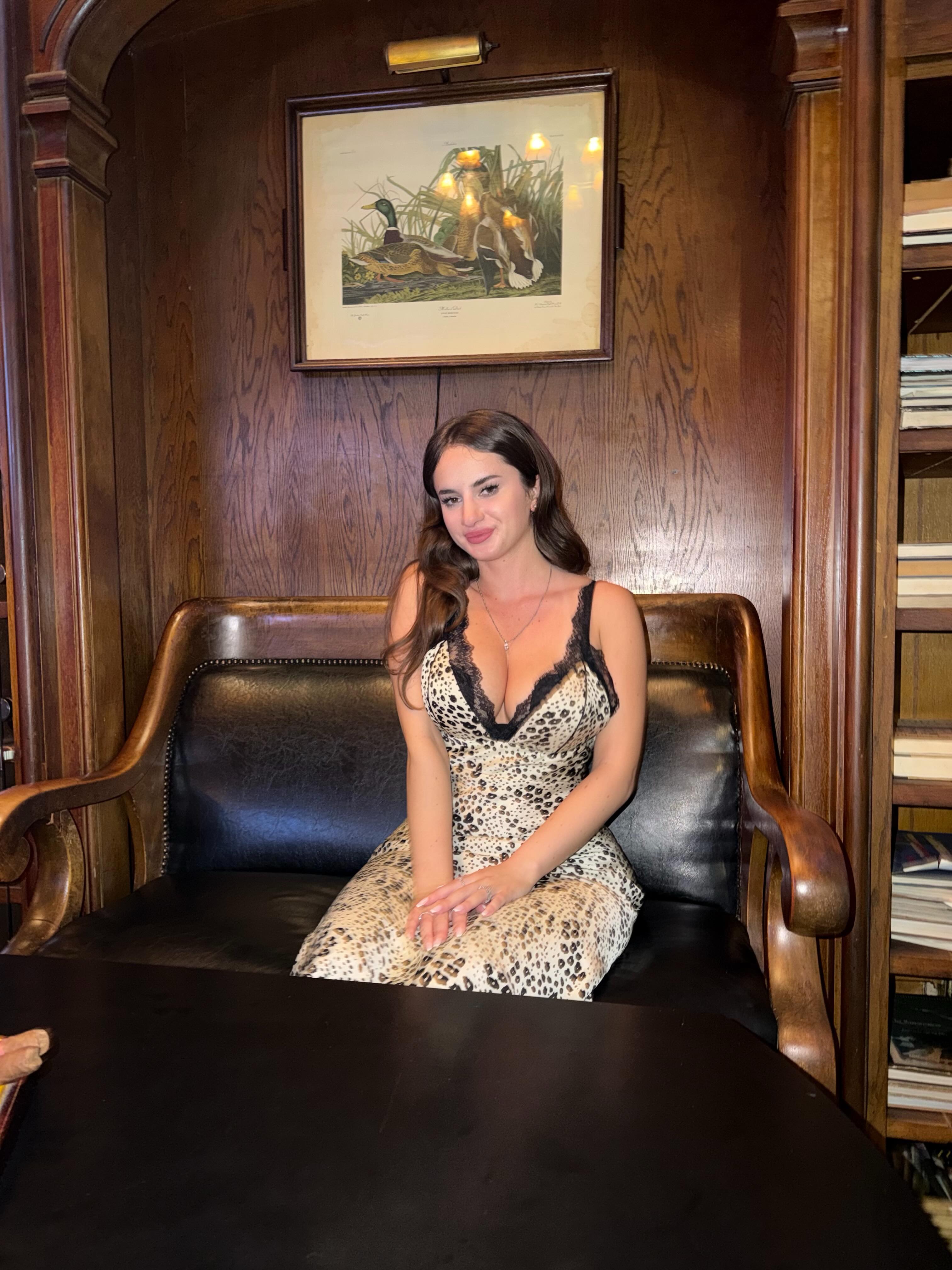 aviebby sitting on a leather bench wearing leopard print dress with black lace in wood paneled room