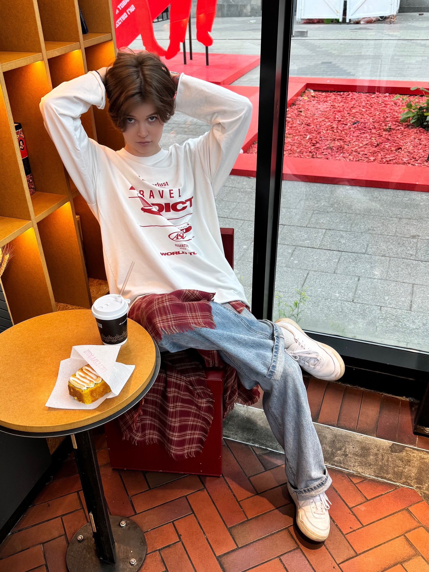 awondrr sitting with hands behind head wearing white long-sleeve graphic shirt in cafe interior by window