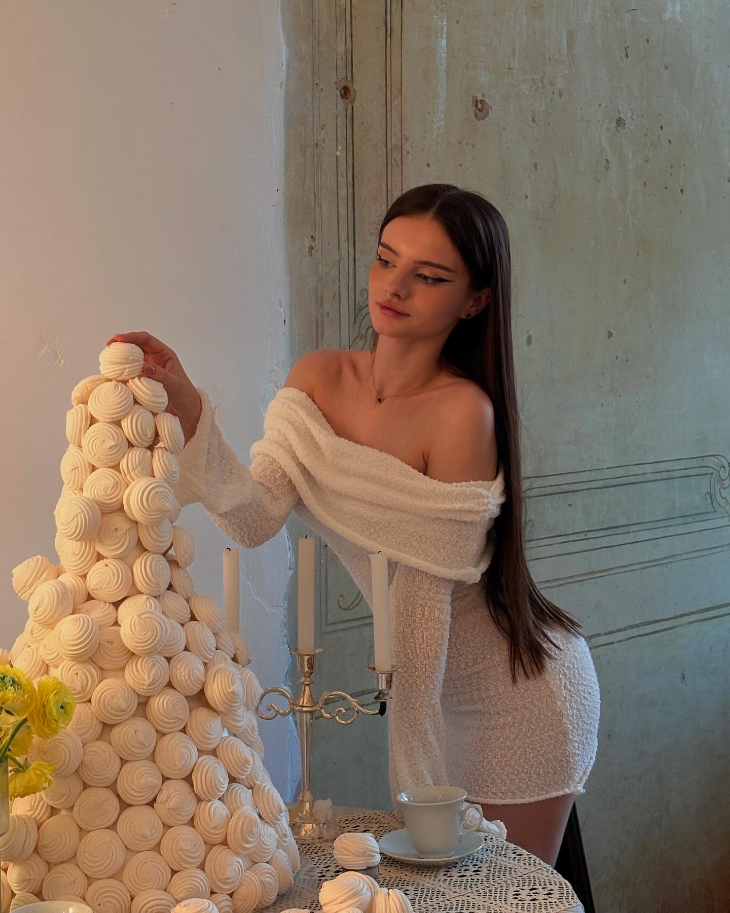 bellacle posing beside a meringue tower wearing white off-the-shoulder mini dress in room