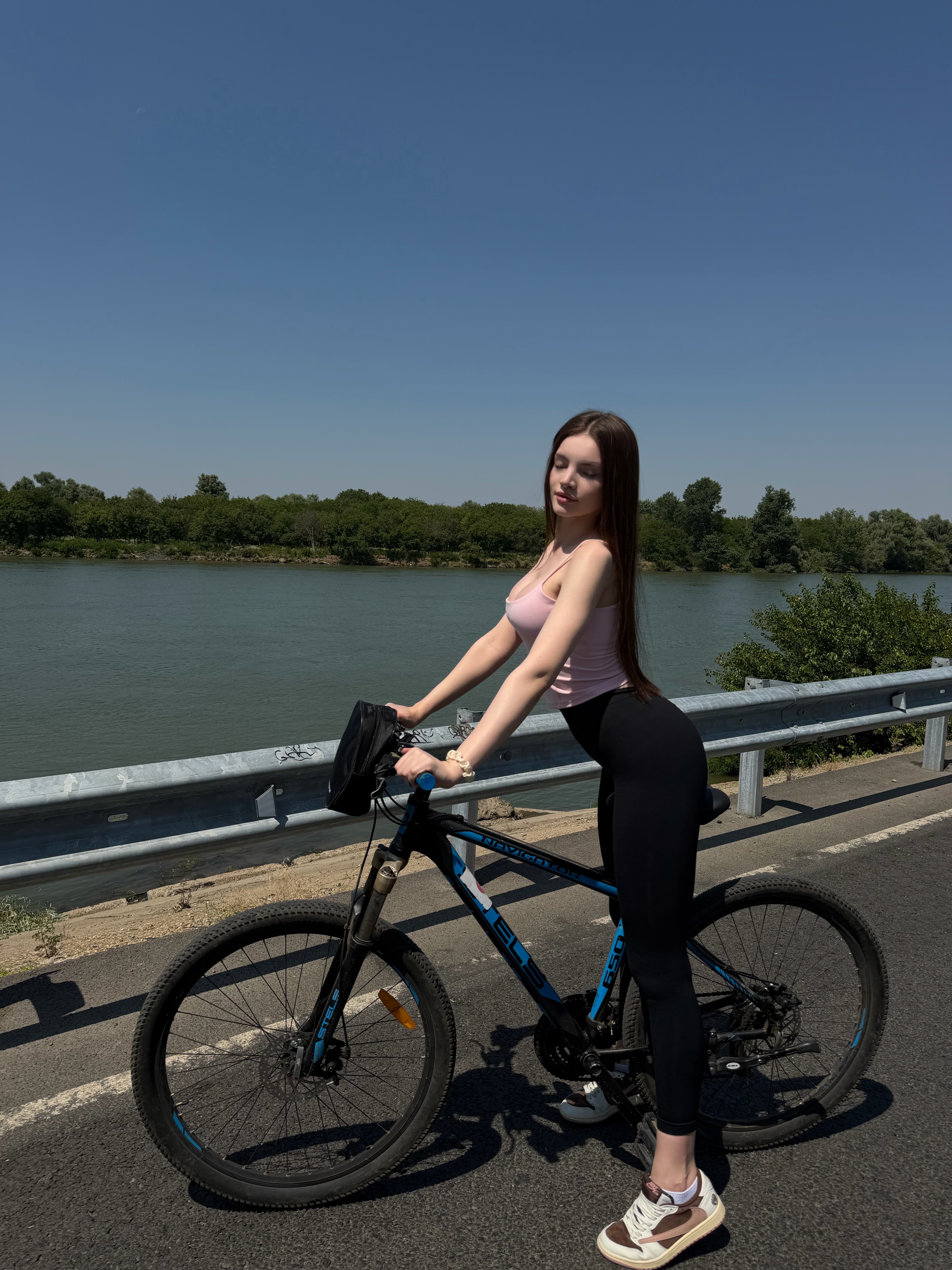 bellacle standing with a bicycle wearing pink tank top in road beside river