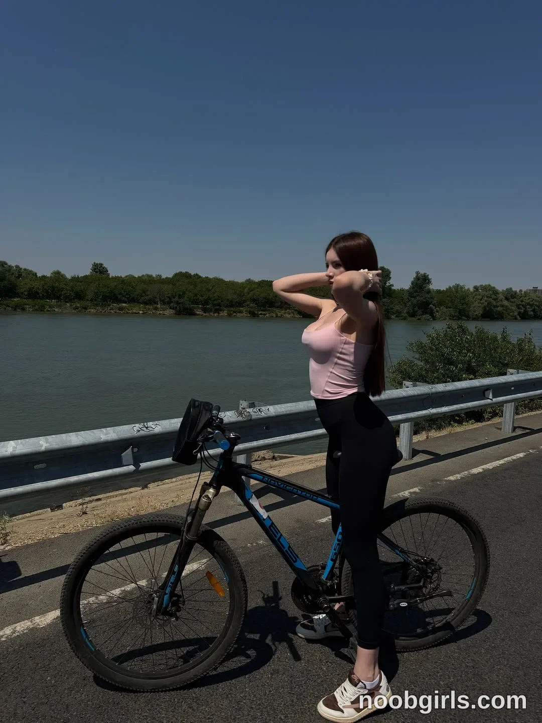 bellacle straddling a bicycle with hands raised behind head adjusting hair wearing light pink camisole tank top