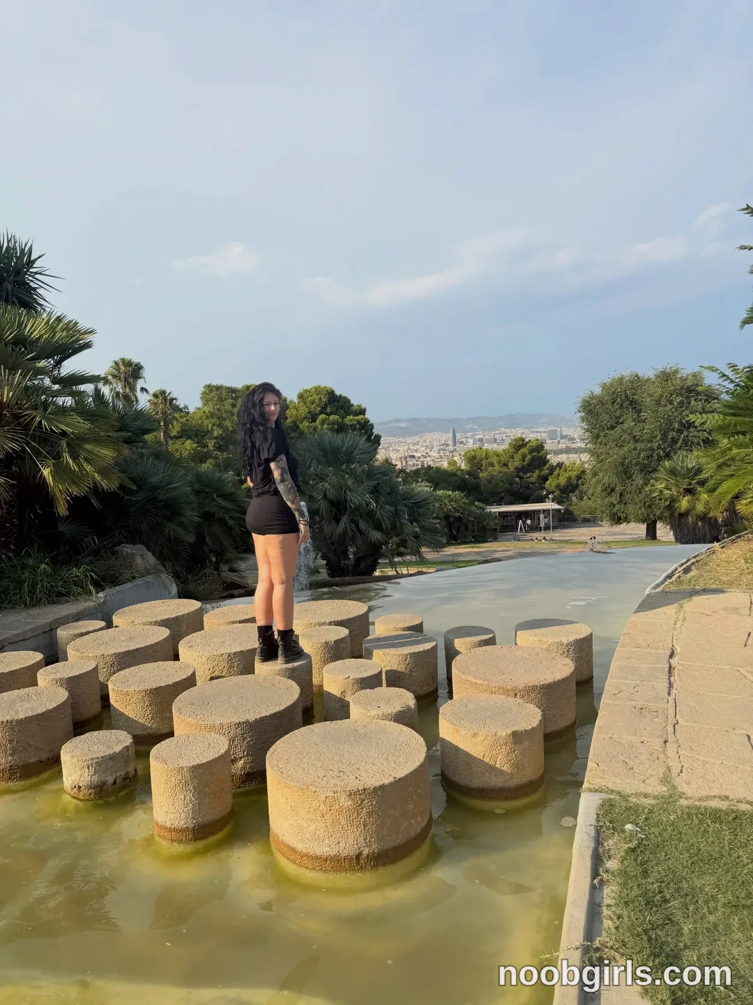 bexbex945874 standing on stepping stones wearing black t-shirt in park fountain overlooking city at Barcelona