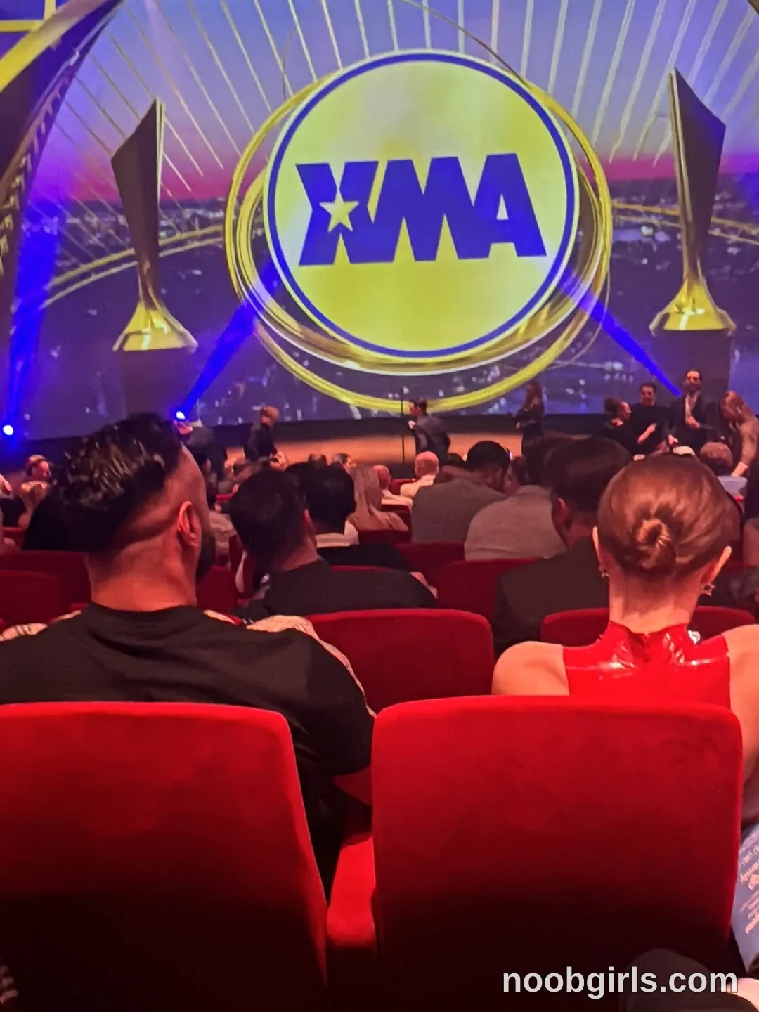 BunnyBlondyX sitting with her back to the camera wearing red sleeveless high-neck garment in auditorium
