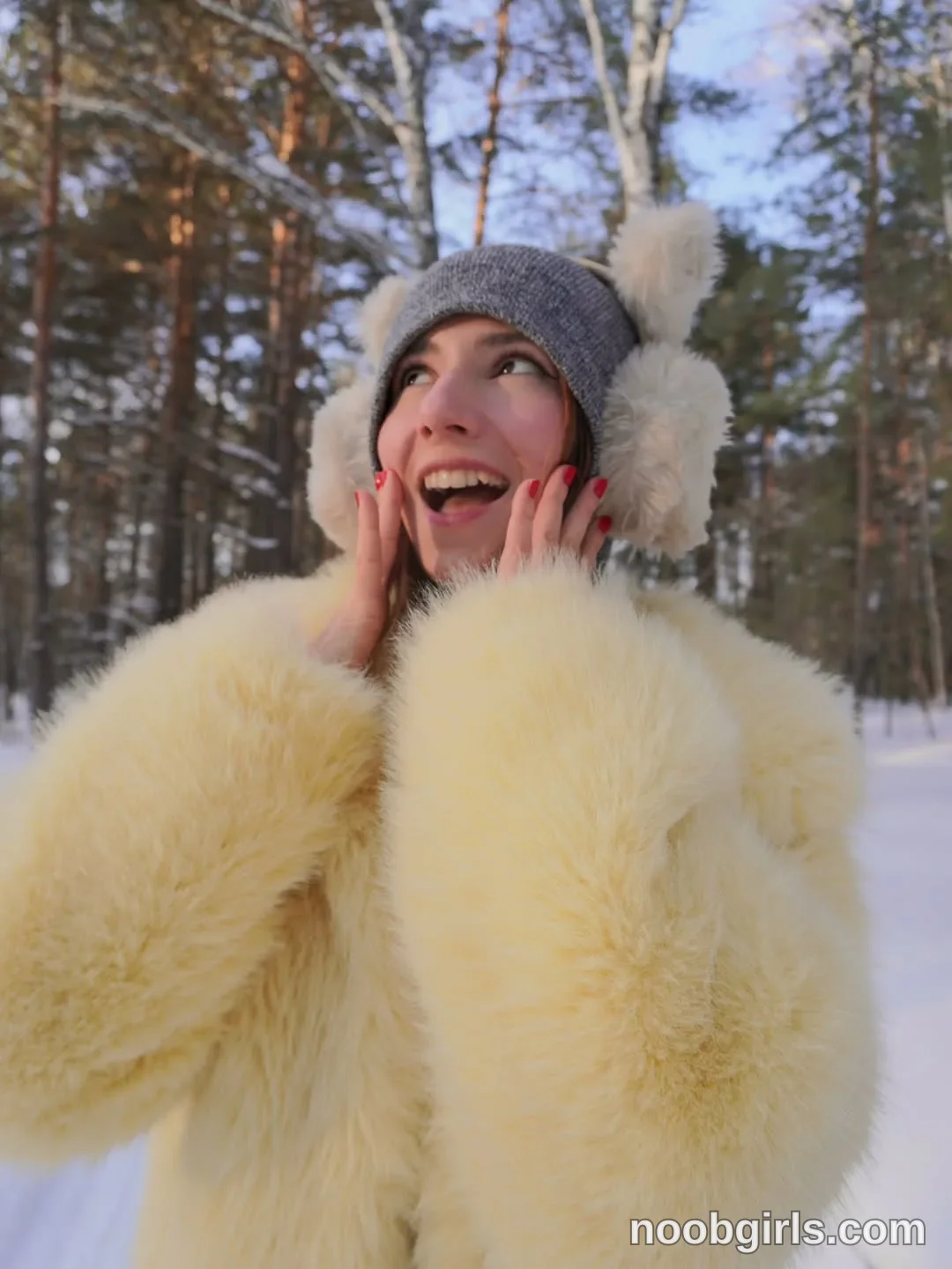 Maria touching both cheeks with her hands wearing gray knitted hat with white furry pom-poms in snowy forest