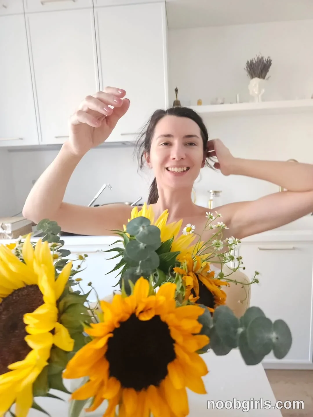 Maria touching her hair with one hand wearing light-colored sleeveless top in kitchen