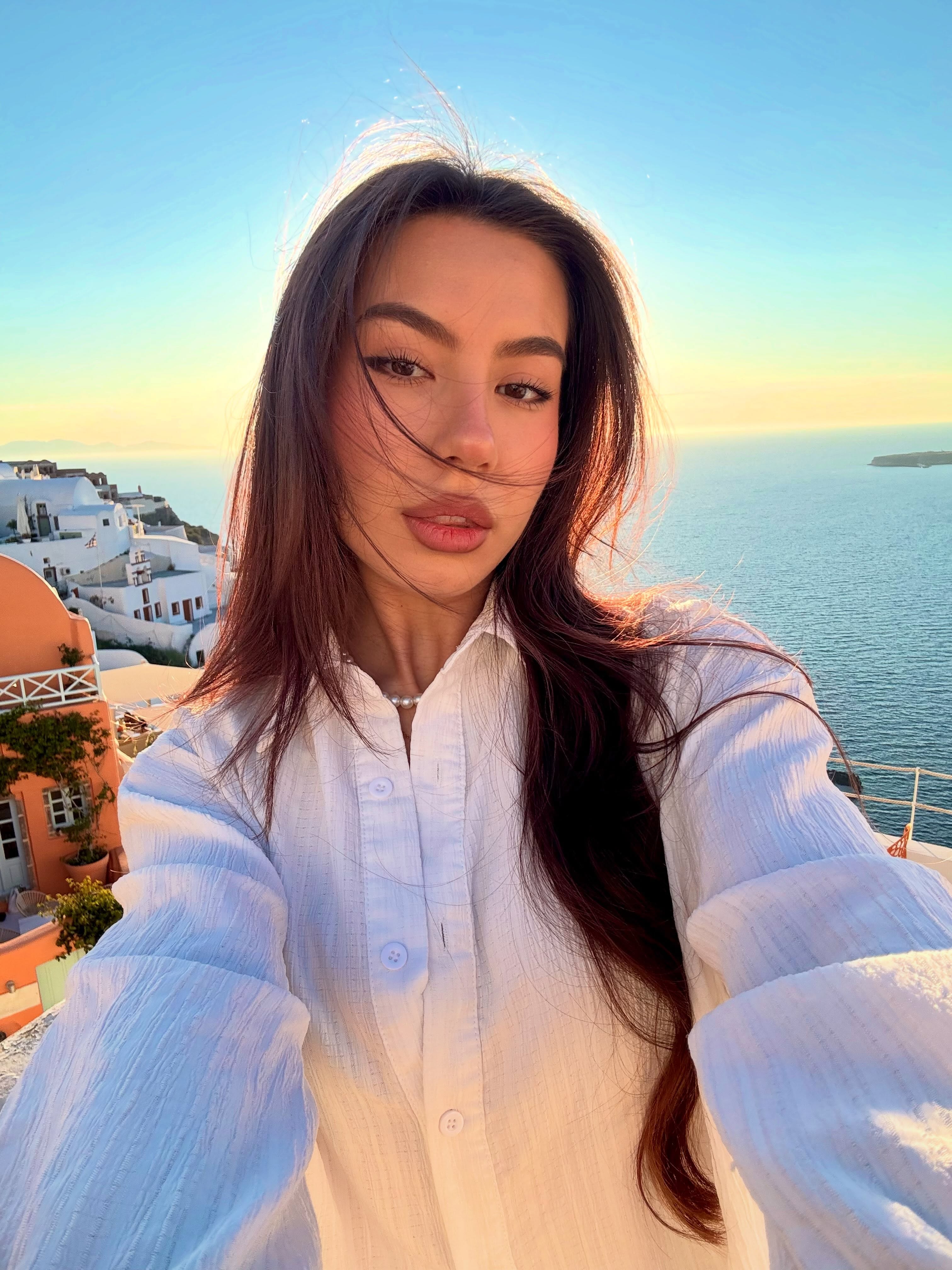 deniseglows taking a selfie wearing white button-up shirt in cliffside village overlooking the sea at Santorini