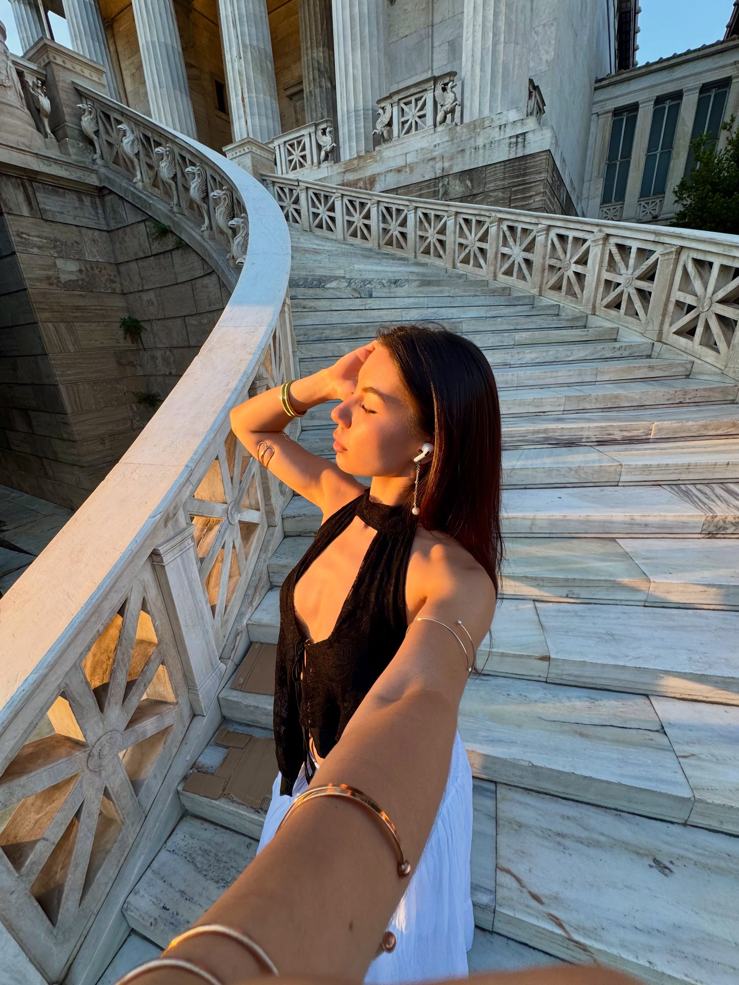 deniseglows taking a selfie with eyes closed wearing black halter top in marble staircase outside classical building