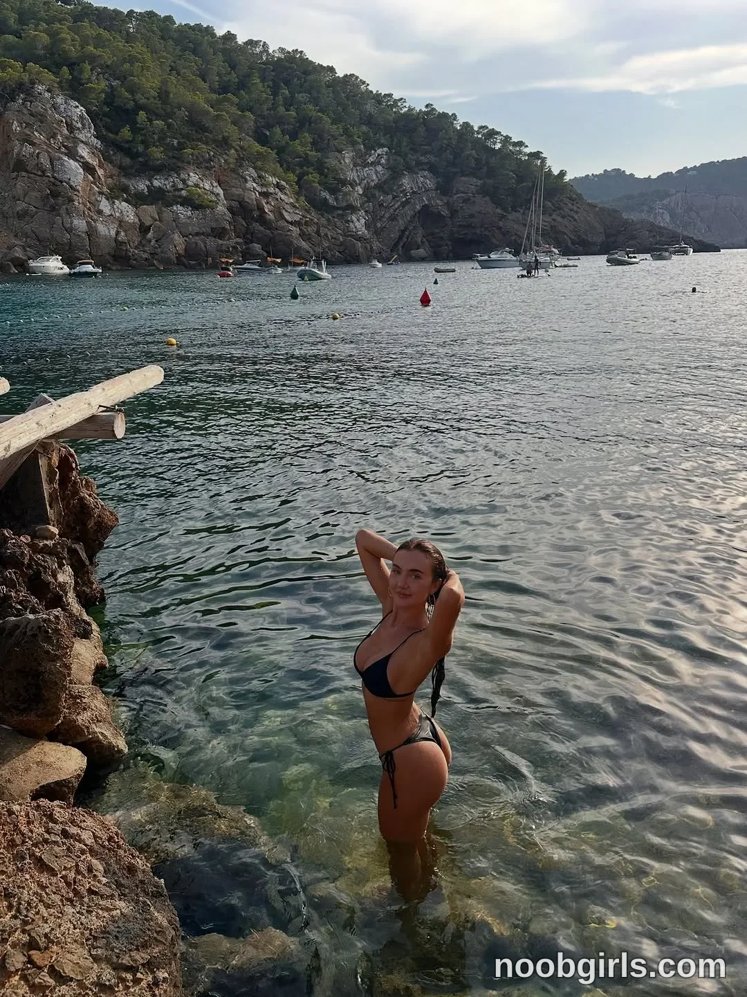 Lilah standing in shallow sea water with hands behind head wearing black bikini top in rocky cove or bay in Ibiza, Spain