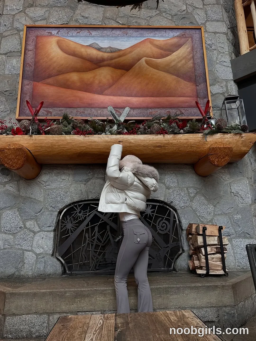 Lilah standing with back to camera wearing white hooded puffer jacket with fur trim on hood in front of a stone fireplace