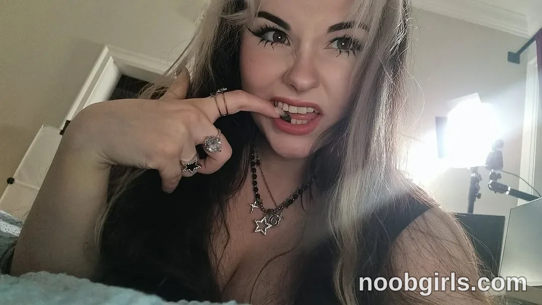 MissTery Leaning toward the camera with one index finger against her front teeth wearing Black sleeveless top in room