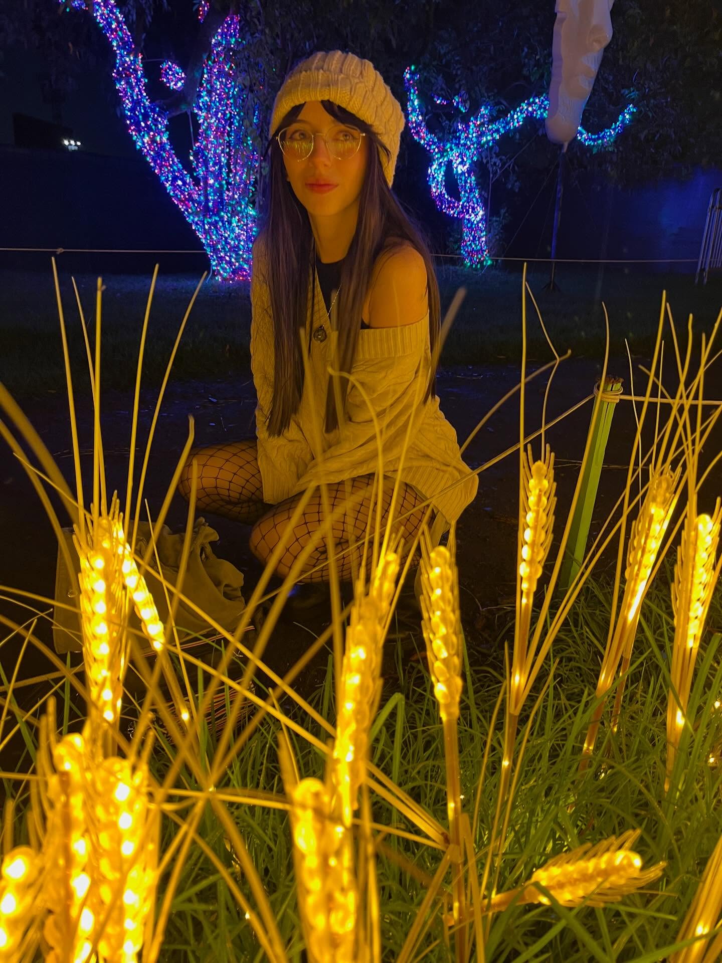 Jelly Crouching among the illuminated stalks with her knees bent wearing Cream ribbed knit beanie in planted area