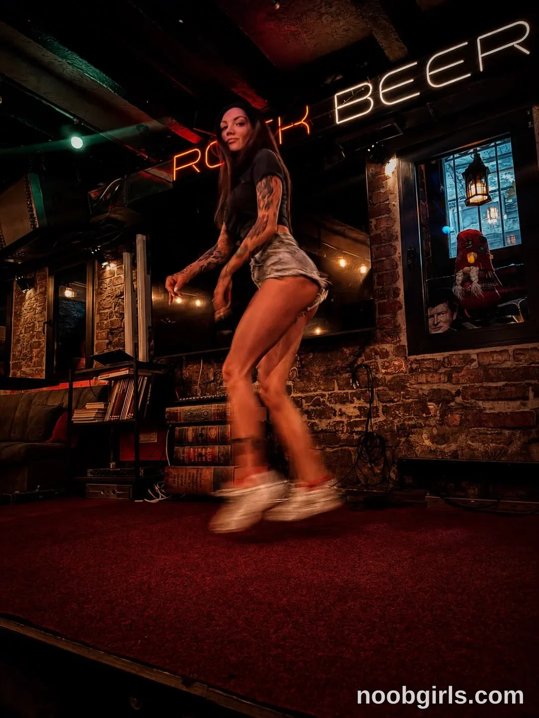 Alice Standing with bent knees wearing Black short-sleeved top on a carpeted platform in a bar