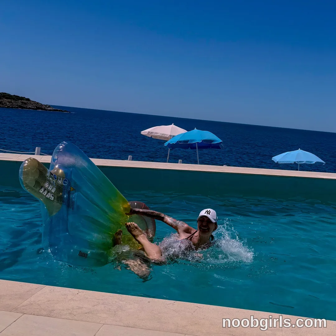 Alice partly submerged in the pool wearing White baseball cap with a black NY logo in swimming pool beside the sea