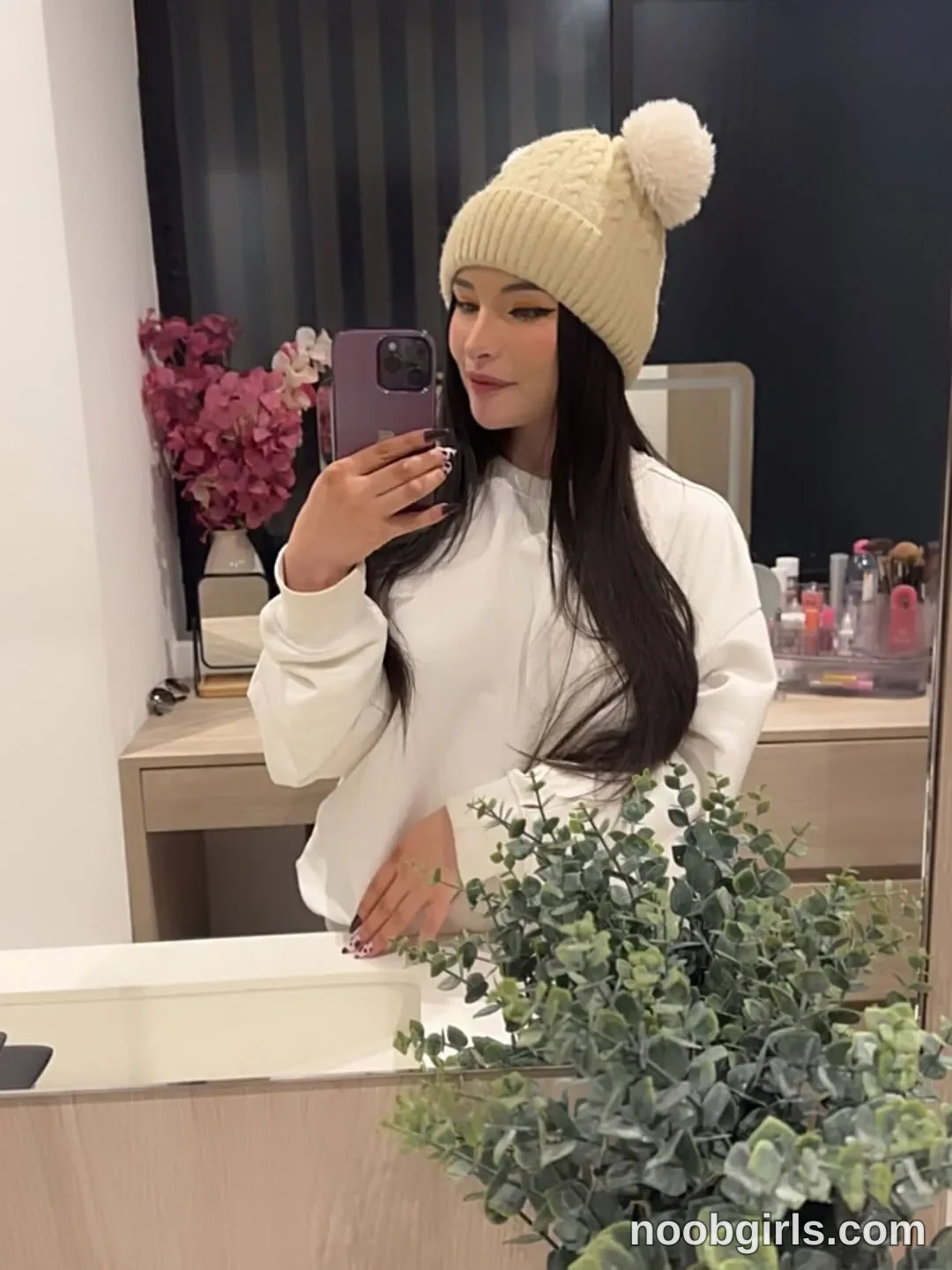 lau__1 holding a smartphone toward the mirror with one hand wearing cream cable-knit beanie with a pom-pom