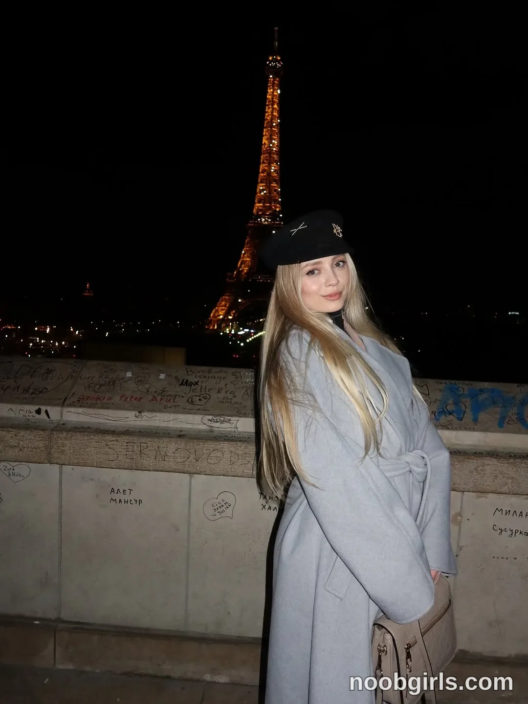Nastya standing with her body angled sideways wearing light grey belted coat beside a graffiti-marked stone parapet