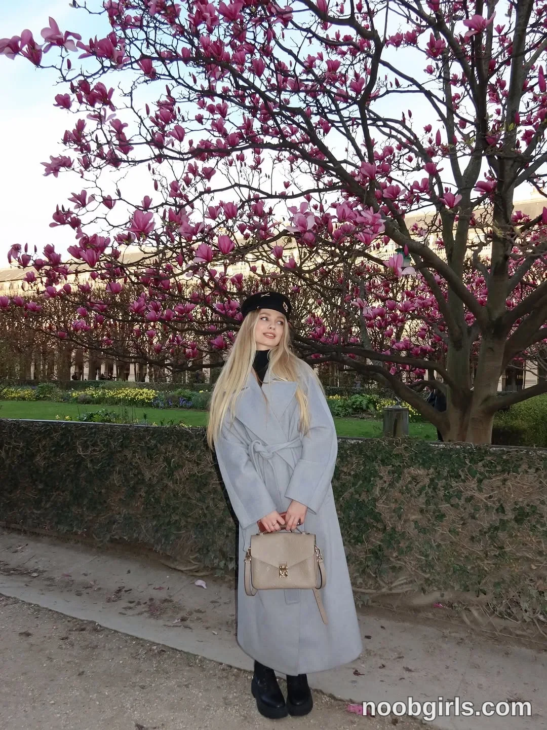 Nastya standing on the path wearing long light grey coat with a tied waist belt in garden