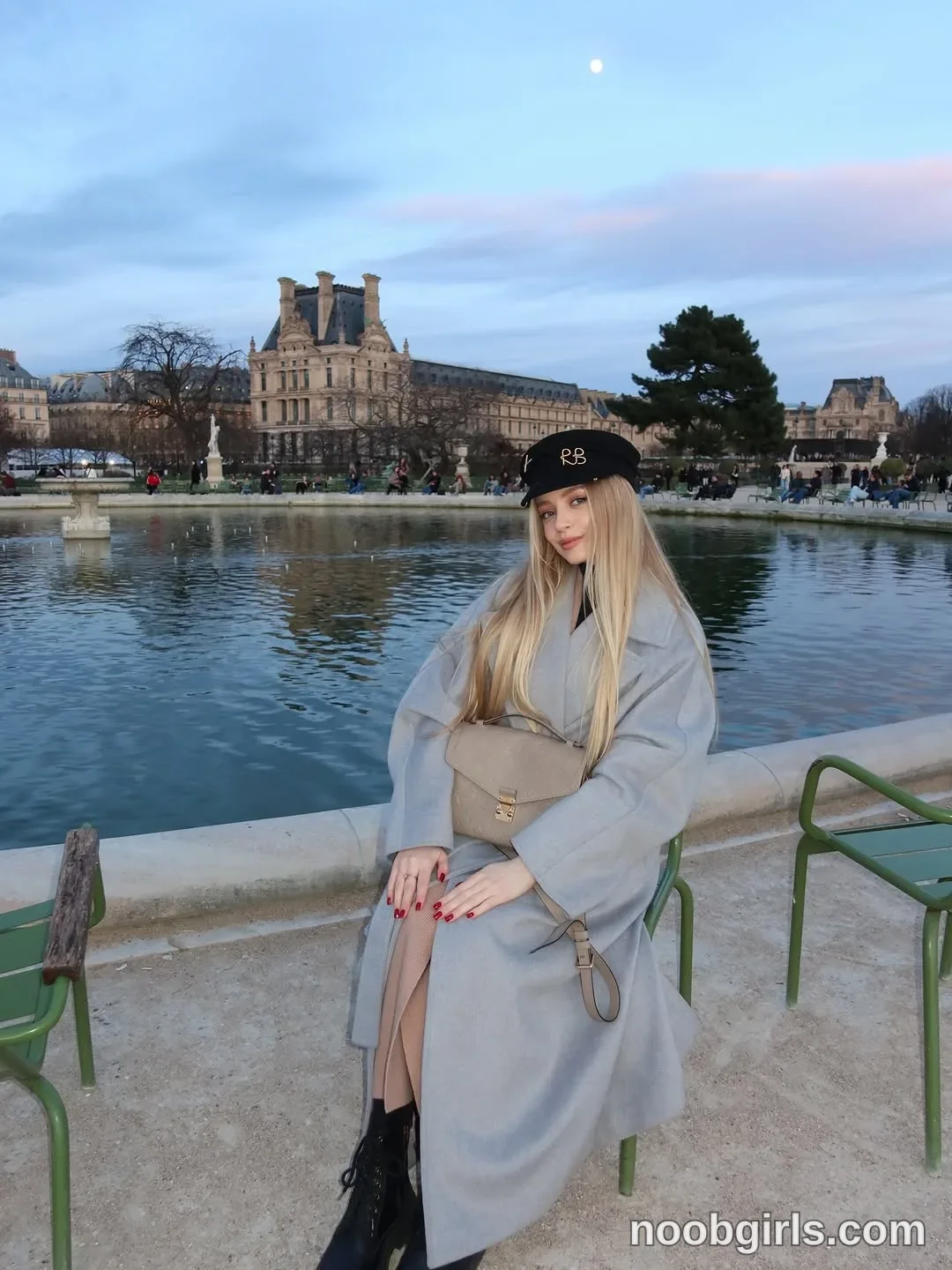 Nastya sitting on a green metal chair with her hands resting on her lap wearing Long light-grey coat