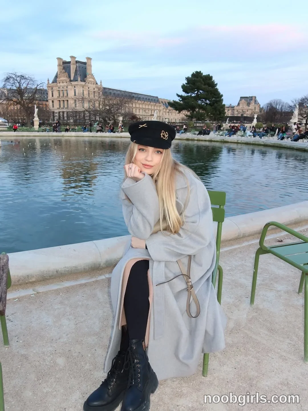 Nastya sitting on a green chair with her legs crossed wearing Long light-grey coat in garden beside a large circular pool