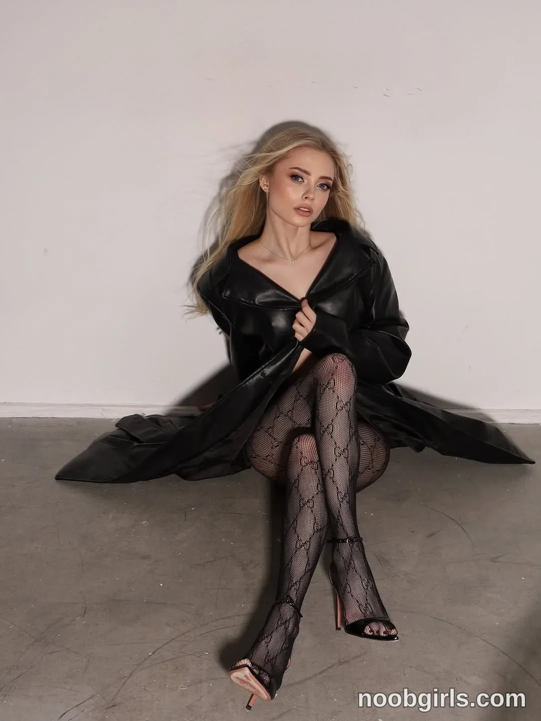 Nastya sitting on the floor with her legs crossed wearing long black leather-look coat in a room