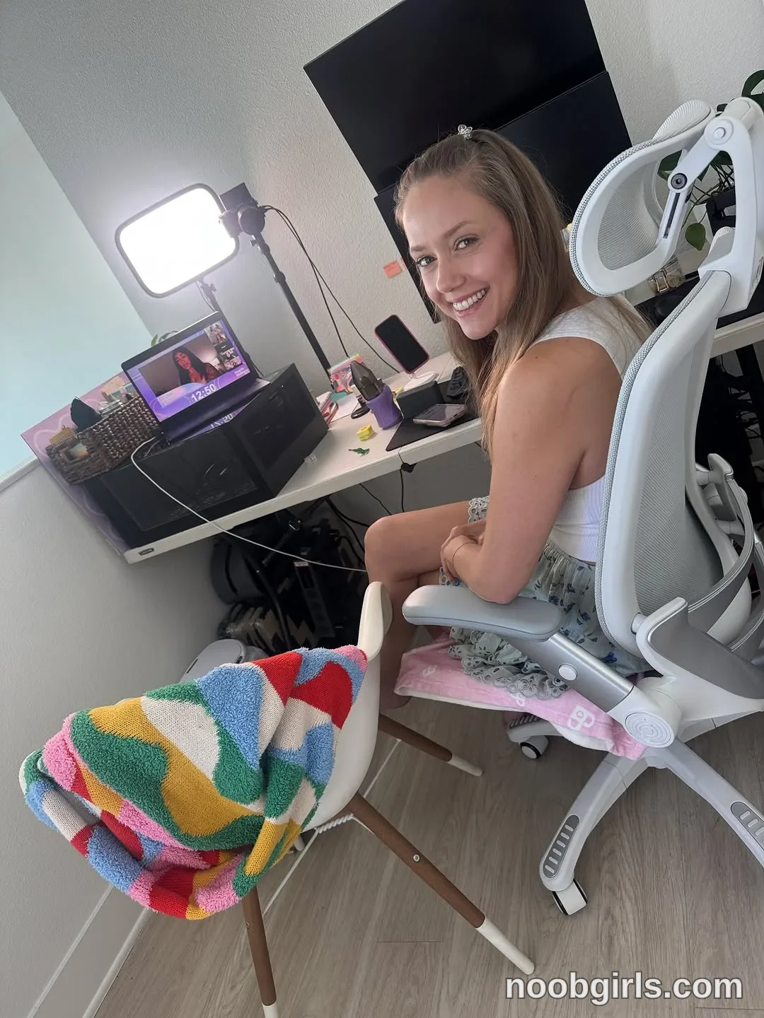 Naked Bakers Sitting sideways in an office chair with her hands near her lap wearing White sleeveless top in room