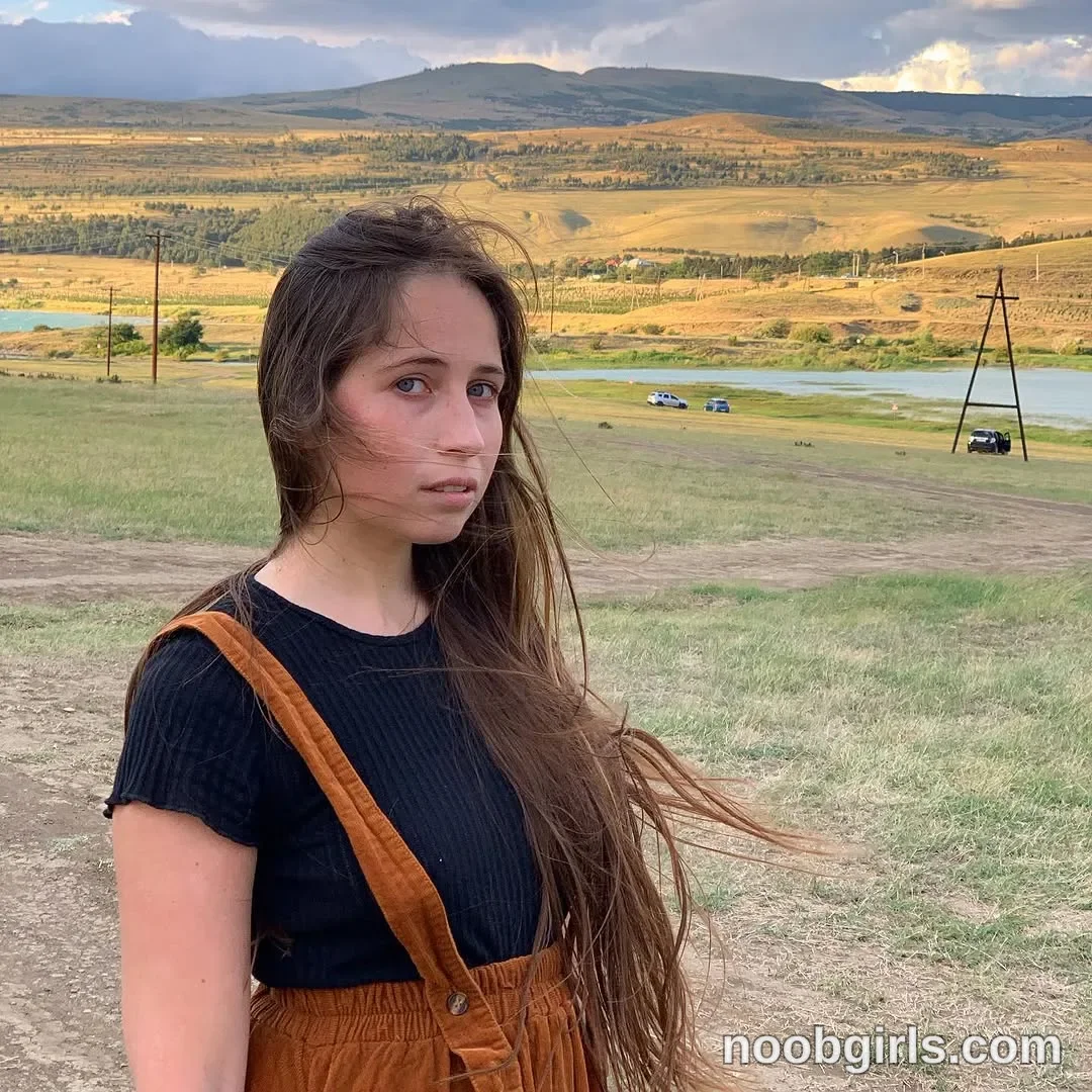 Riley Standing with her body angled sideways wearing Black short-sleeved ribbed top in grassy hillside
