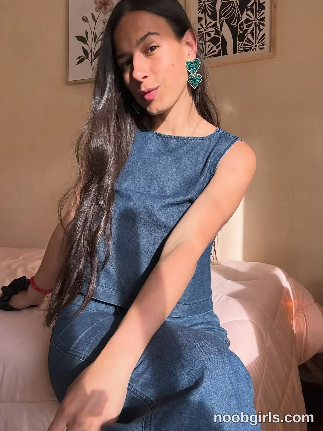 ohanna sitting on a bed leaning toward the camera wearing blue denim sleeveless top in bedroom