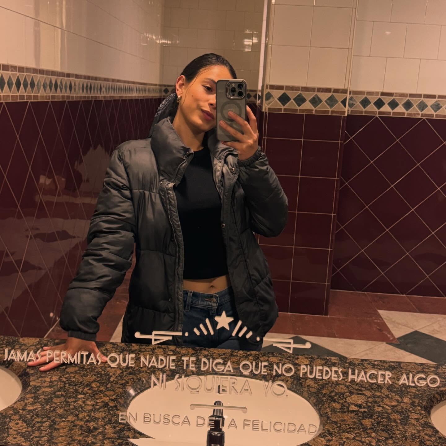 ohanna taking a mirror selfie with a smartphone wearing dark grey puffer jacket in public or commercial restroom
