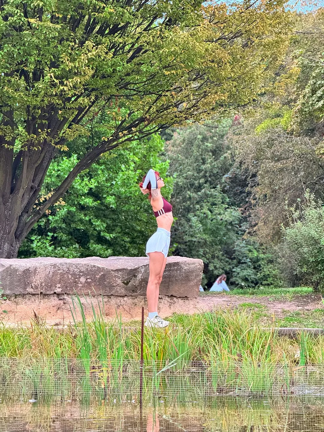 evaoopsy Standing in profile with both arms raised wearing Burgundy sports bra on a grassy bank beside water