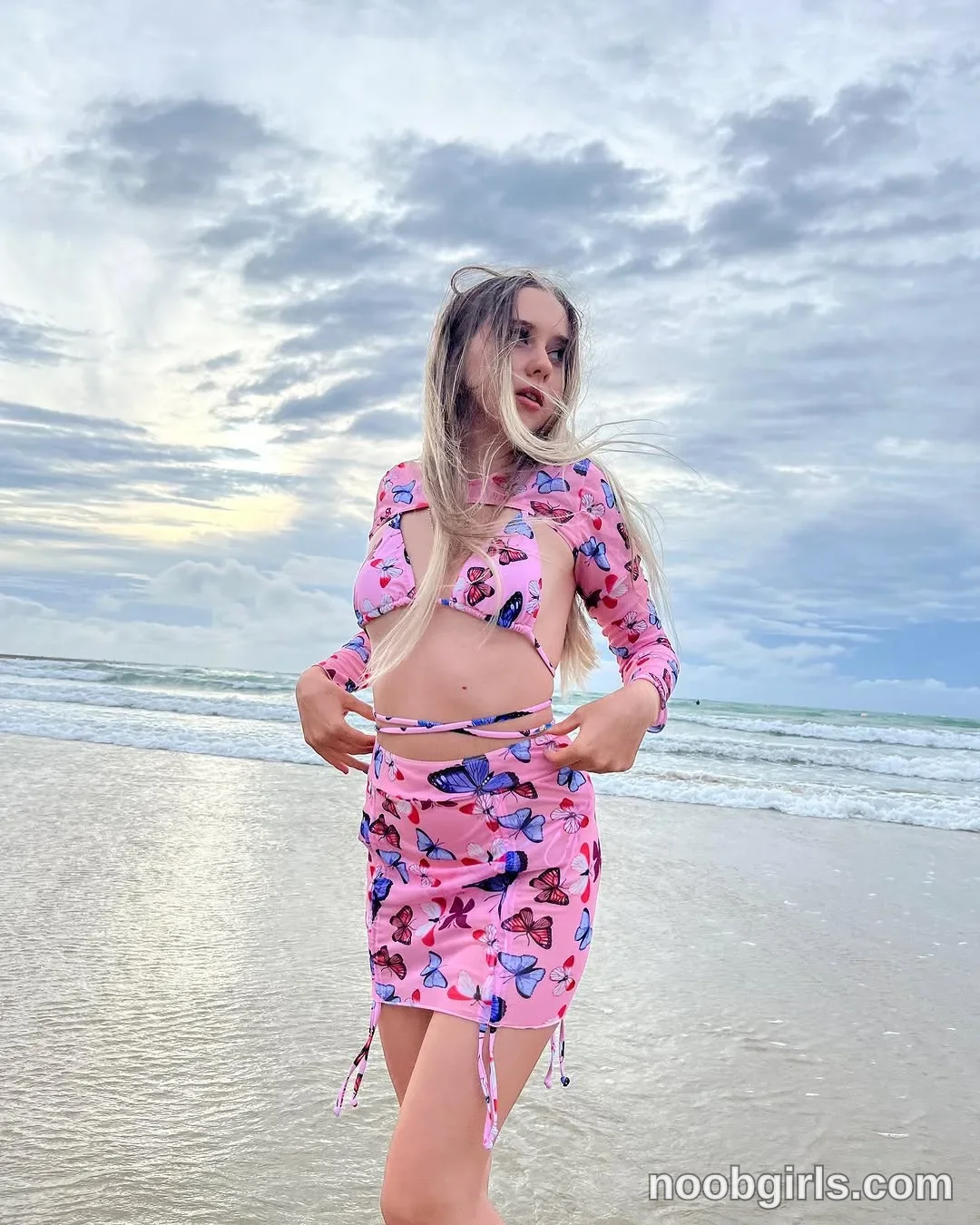 Elina standing on wet sand near the water's edge wearing pink long-sleeve crop shrug with multi-coloured butterfly print