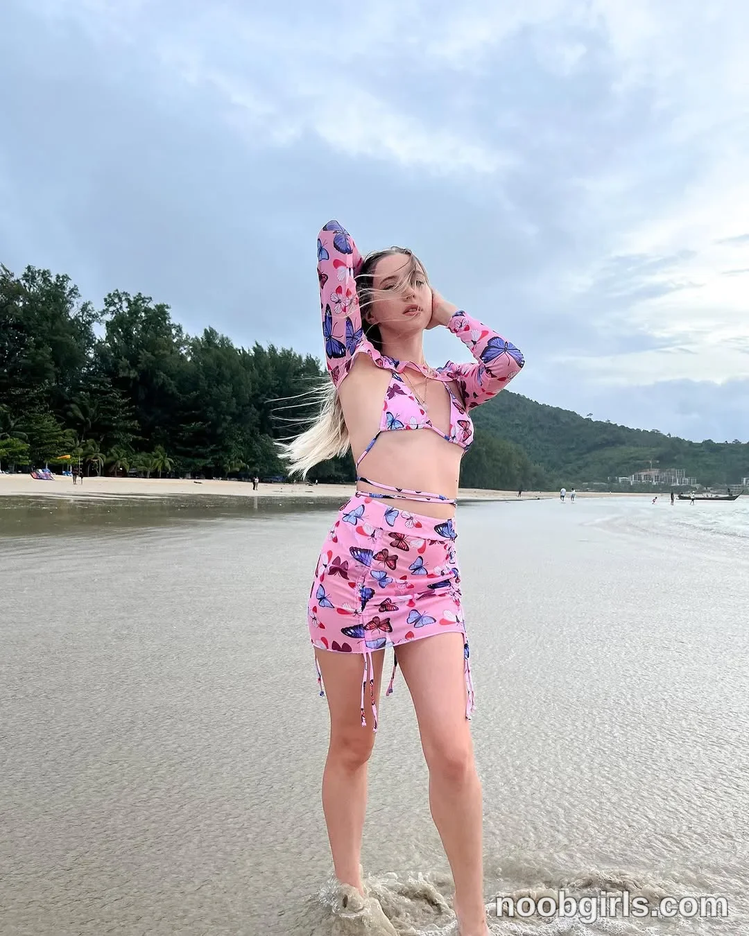 Elina standing barefoot in shallow sea water with both hands raised touching her head
