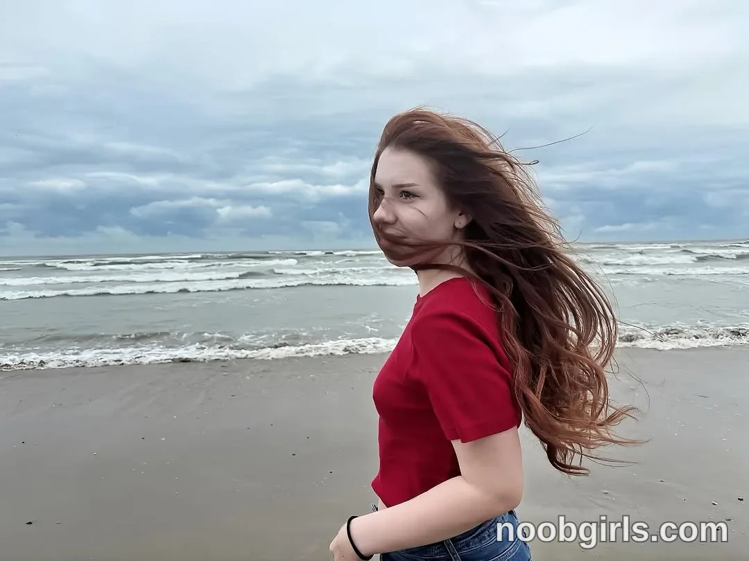 Jo Standing with her body turned sideways wearing Red short-sleeved top on a sandy beach beside the sea