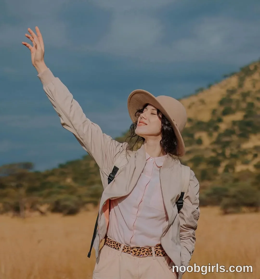 TAANNI Standing with one arm extended upward wearing tan wide-brimmed hat in a field of dry grass